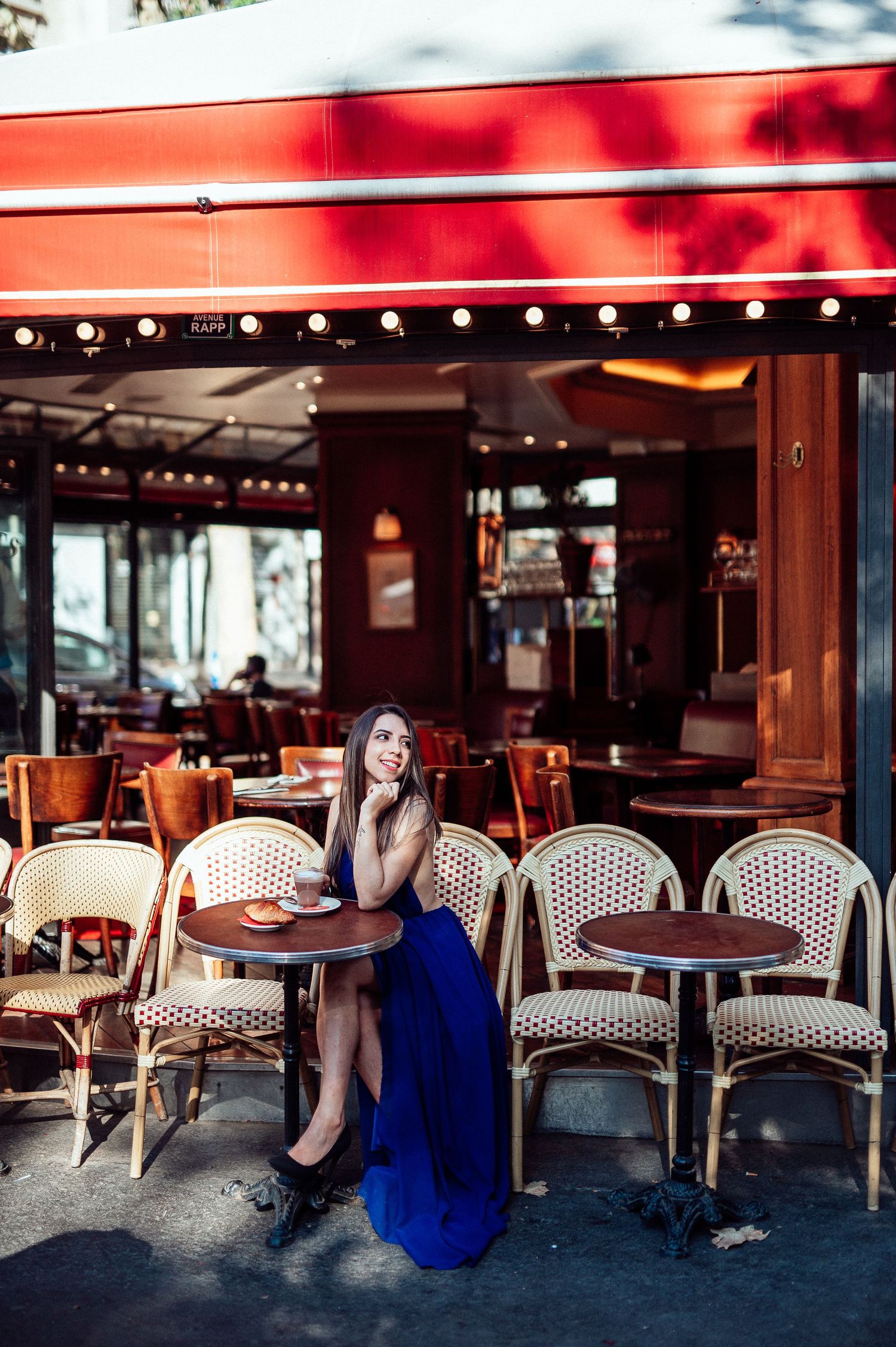 A dreamy Parisian cafe scene with woven chairs and red awning features a person in an elegant blue evening gown.