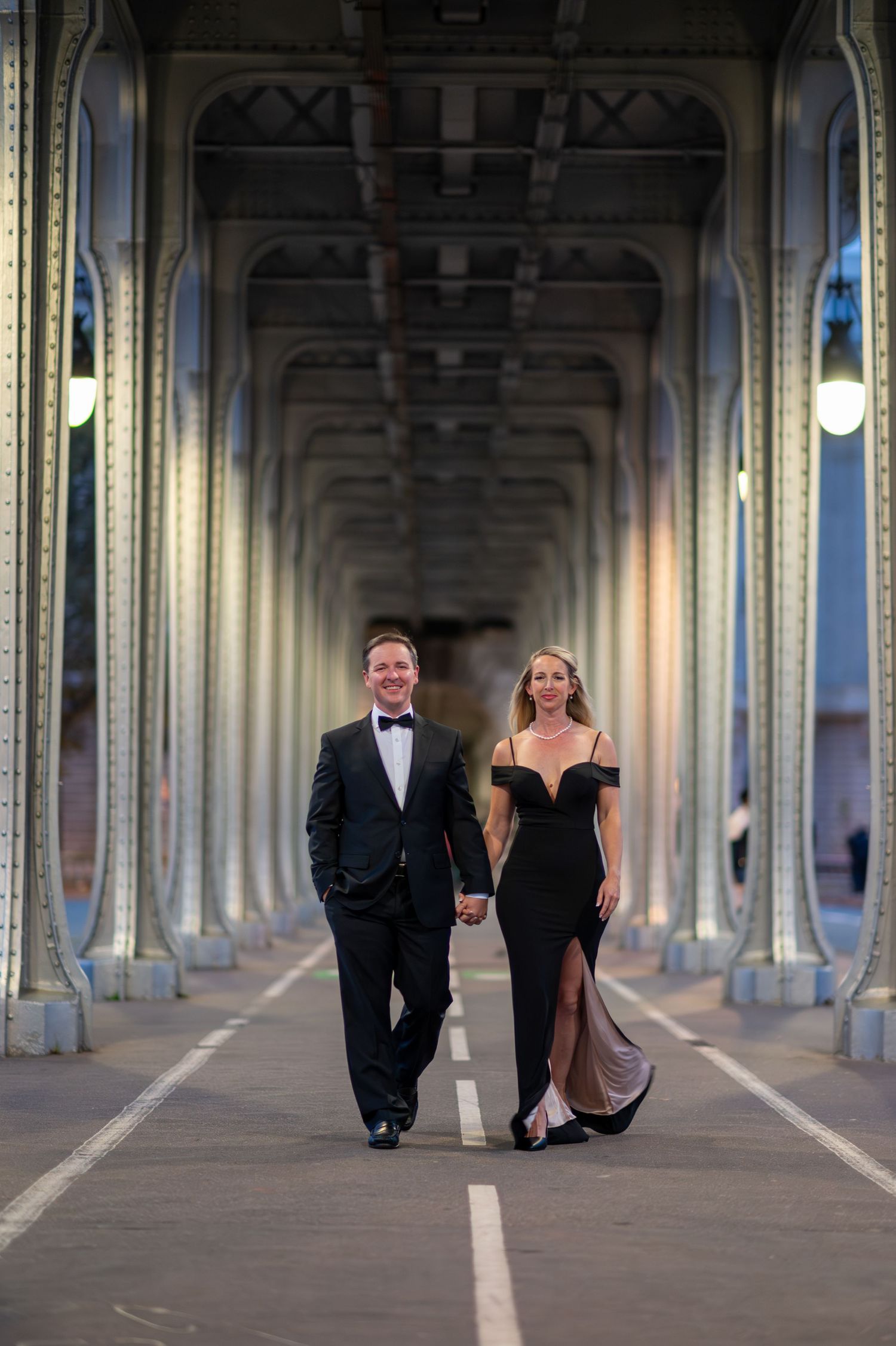 Couple in formal evening wear walk together under the arches of Bir-Hakeim Bridge in Paris at dusk.
