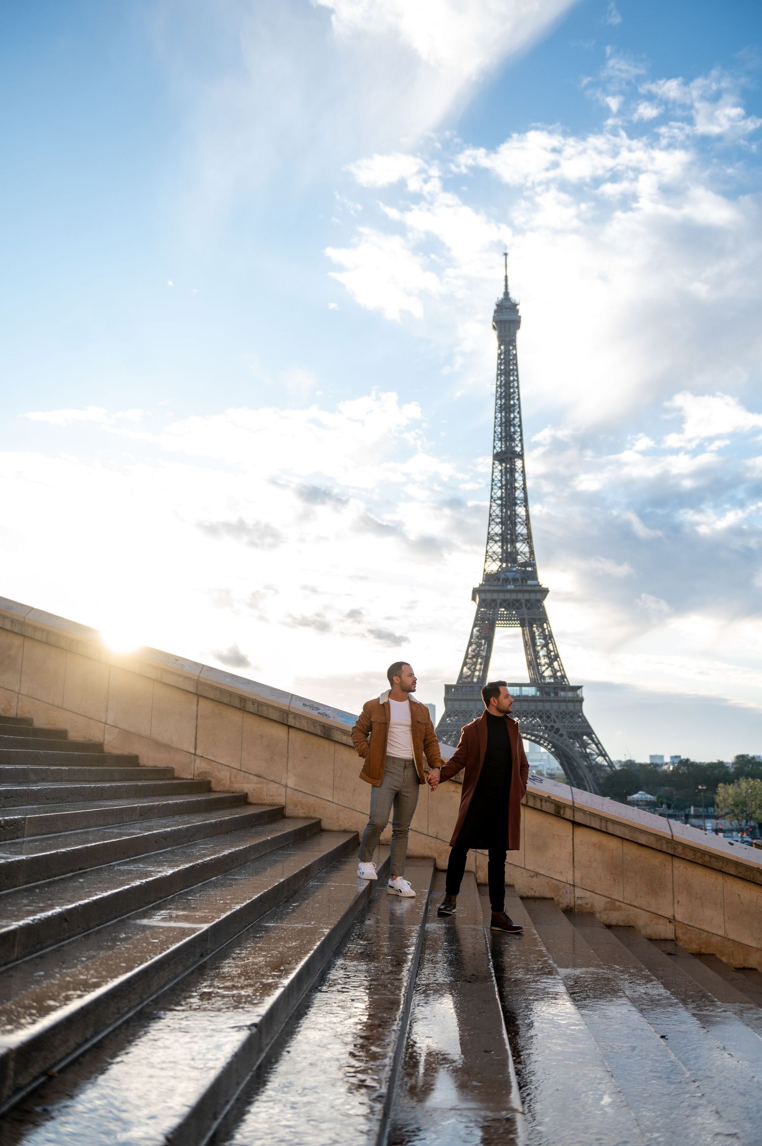 A couple strolls hand in hand on rain-soaked steps near the iconic Eiffel Tower in Paris during golden hour.