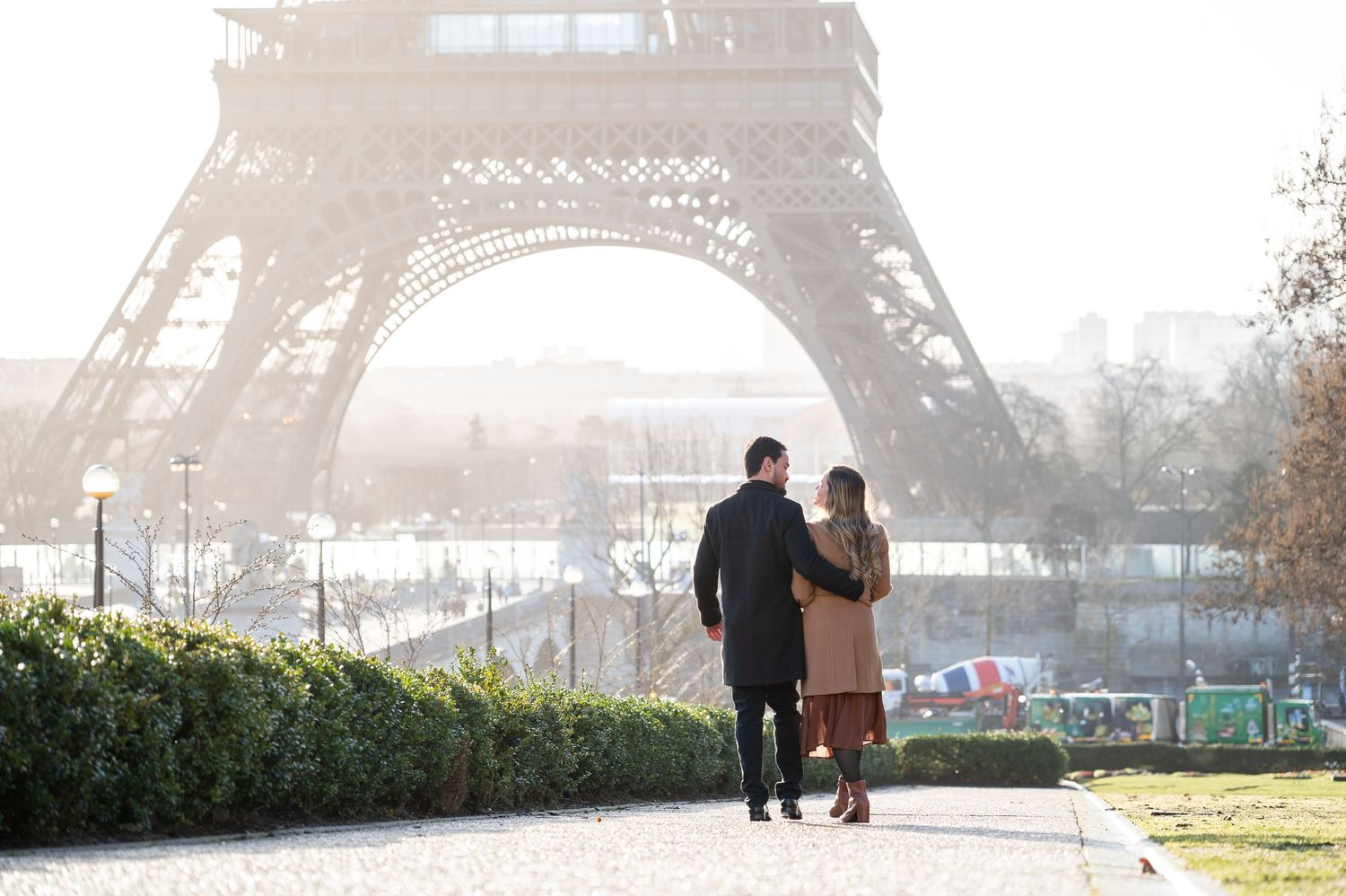 A couple walks hand in hand towards the base of the Eiffel Tower along a garden path.