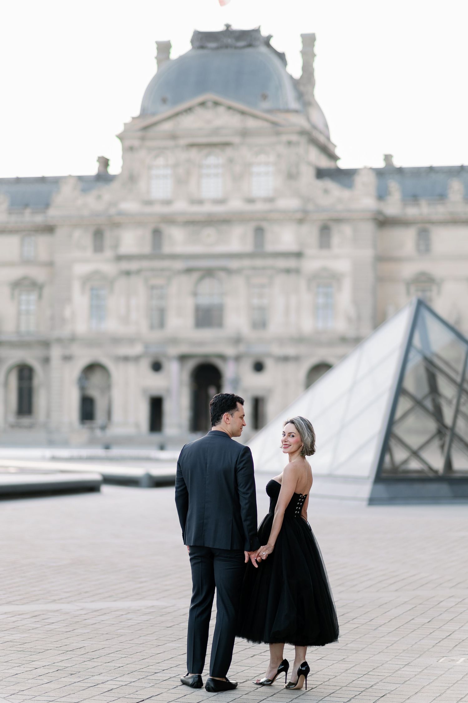 Couple holding hands in front of the Louvre Museum and glass pyramid in Paris, woman in black dress.