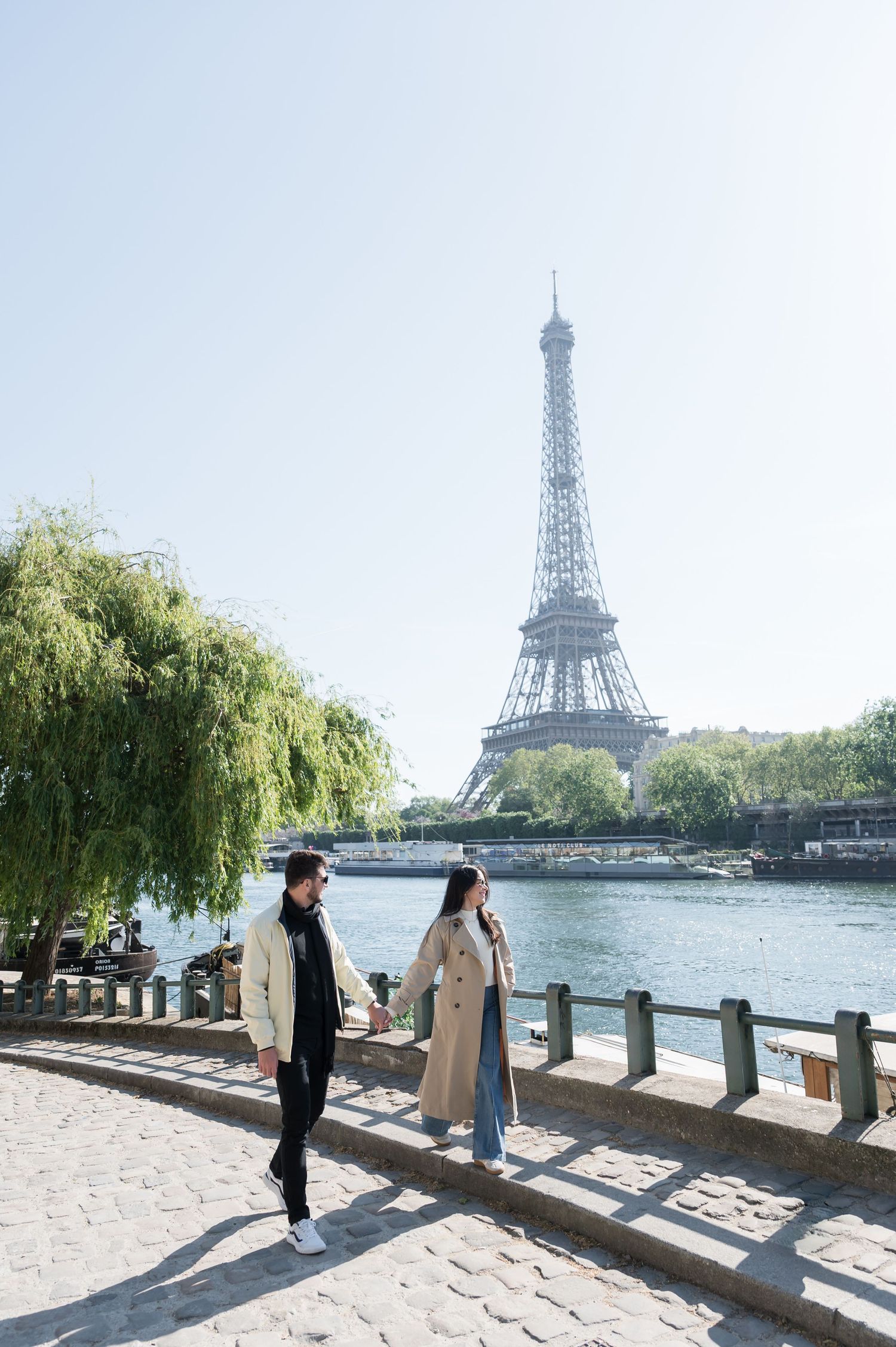 Couple holds hands walking along the Seine River with the Eiffel Tower in Paris, France.
