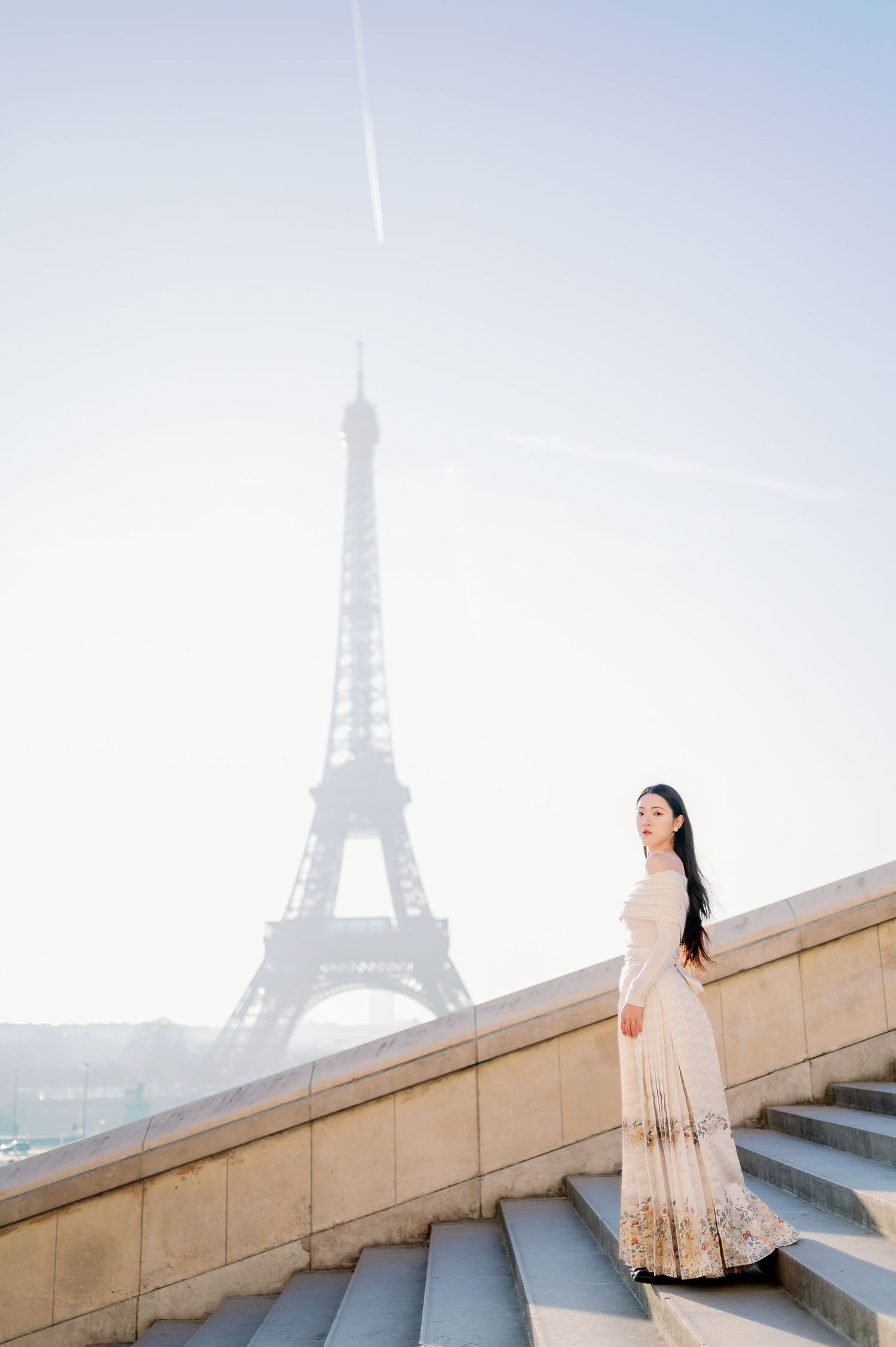 A woman in a cream-colored dress stands on stone steps with the Eiffel Tower visible in the distance.