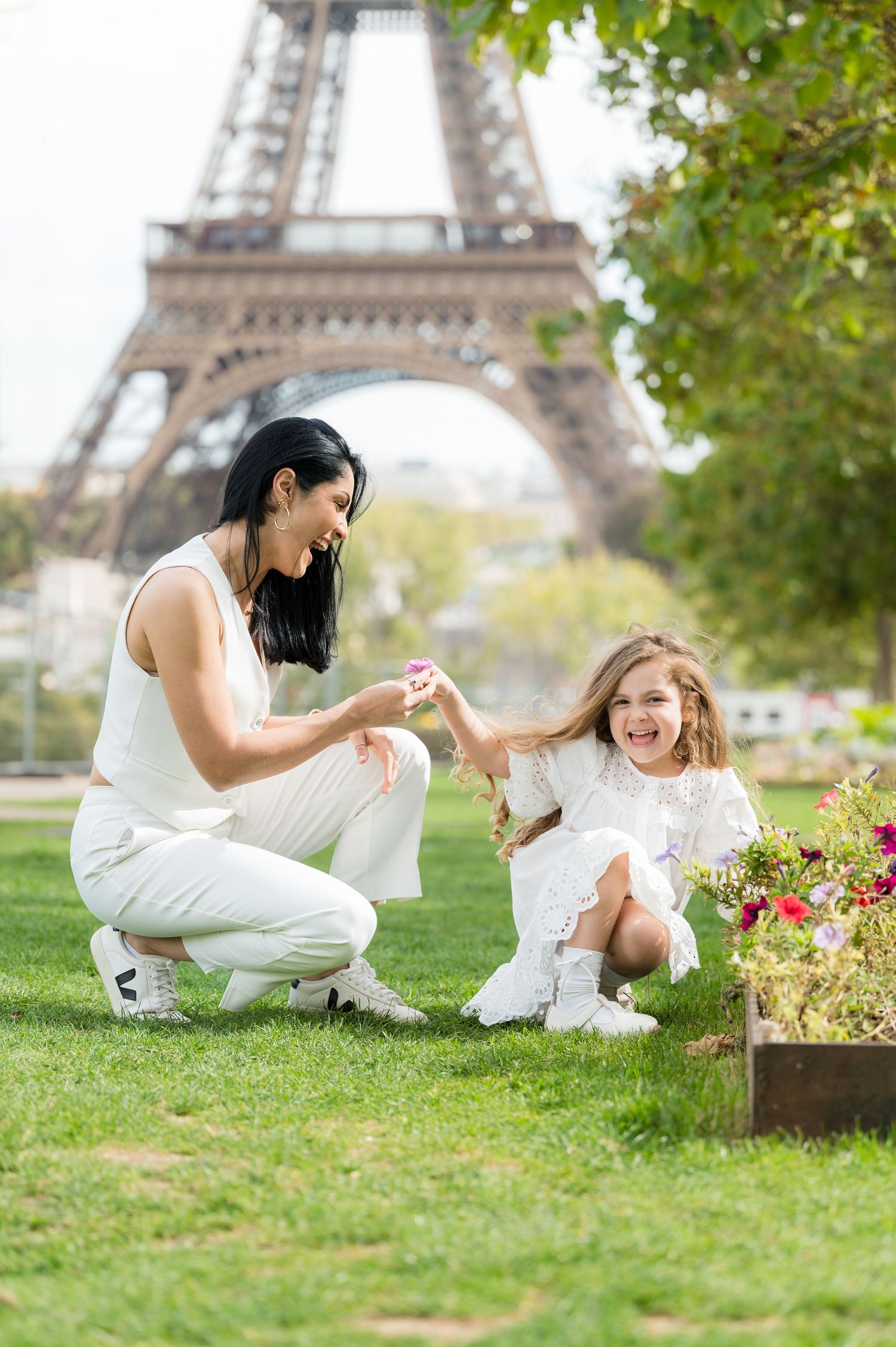 Two people in white outfits share a tender moment on the grass with the iconic Eiffel Tower visible in the background.