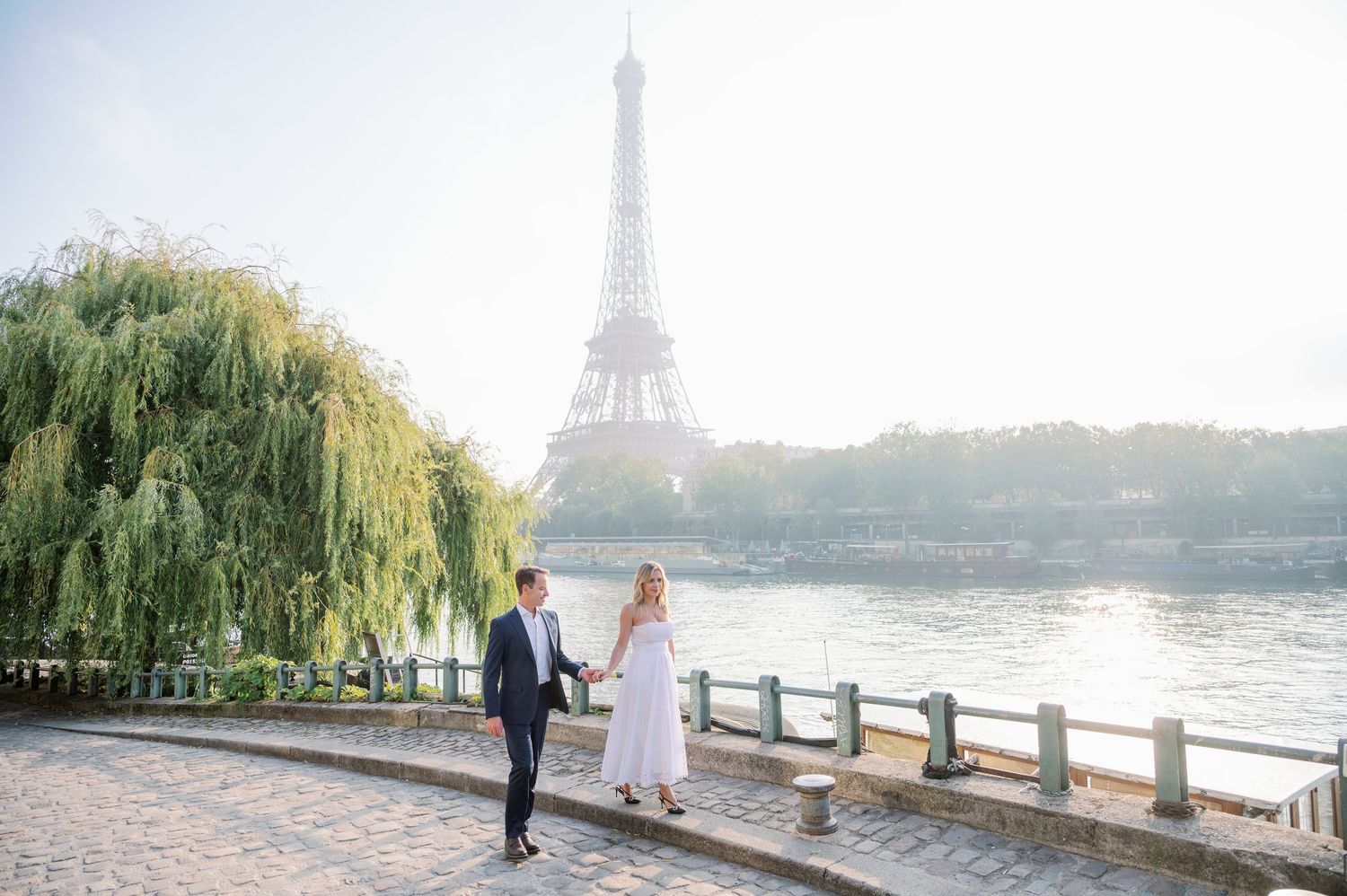 Romantic couple strolls along Seine River with Eiffel Tower and weeping willow trees in soft sunlit Paris backdrop.