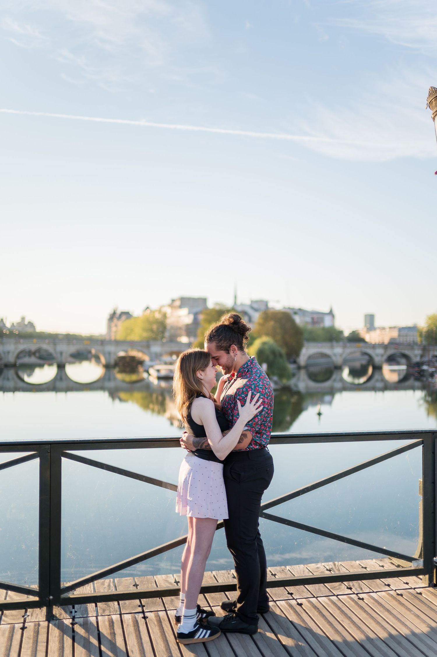 Couple sharing a romantic embrace on a wooden dock with a scenic river bridge and city skyline in the background.