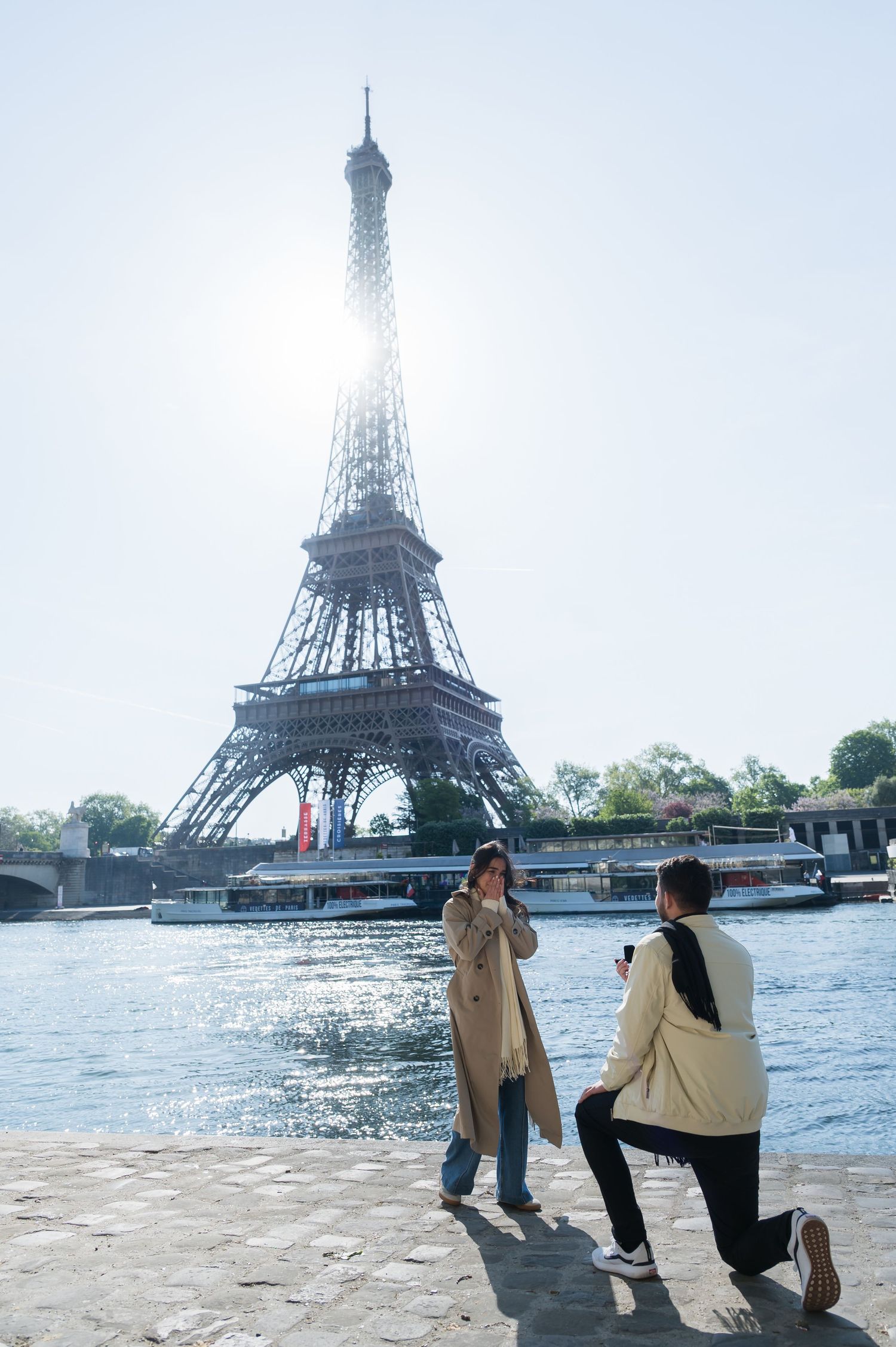 Man proposes to woman on one knee by the Seine River with the Eiffel Tower in Paris, France.