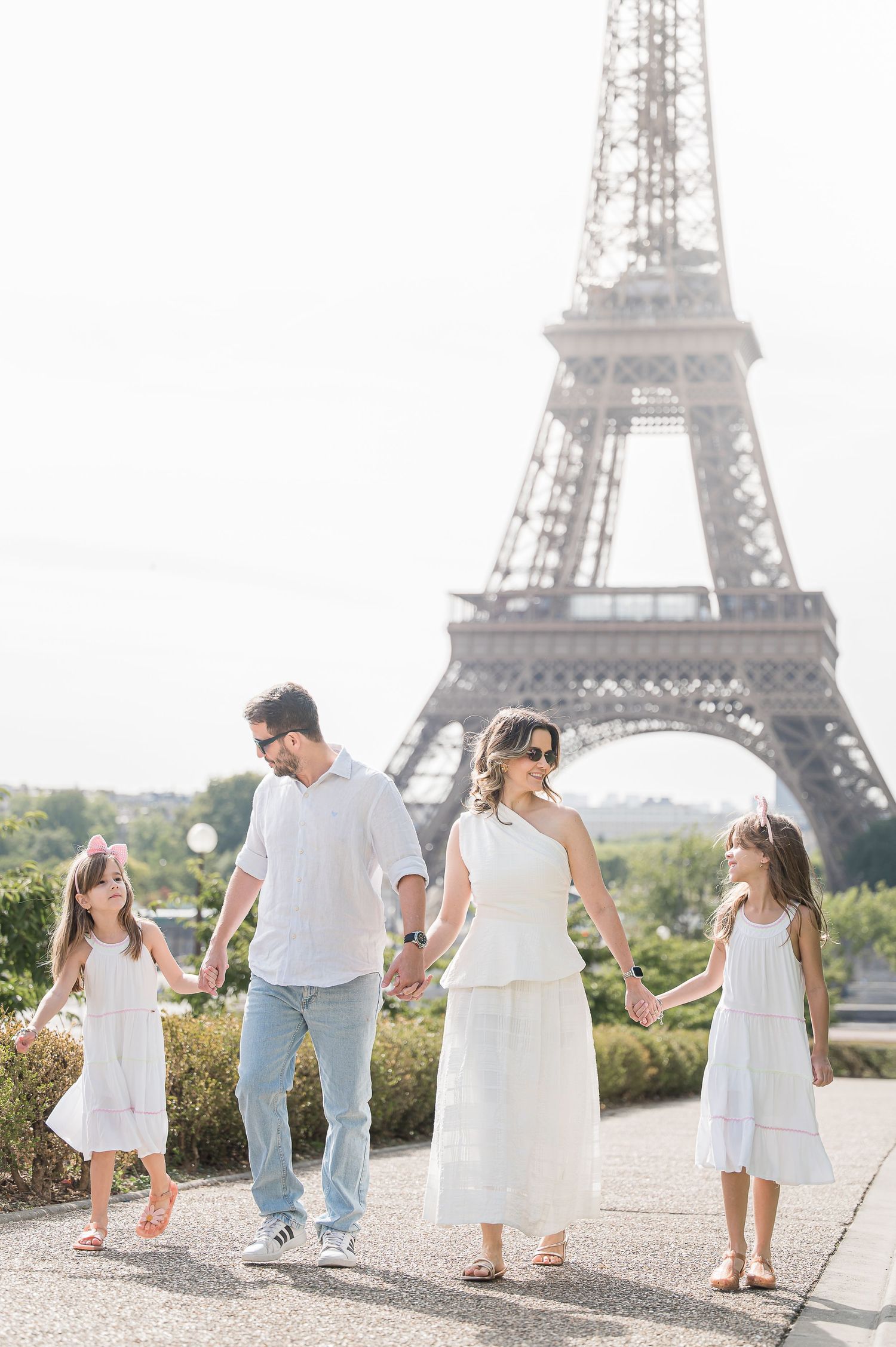 Family of four in white outfits holding hands near the Eiffel Tower in Paris on a bright sunny day.