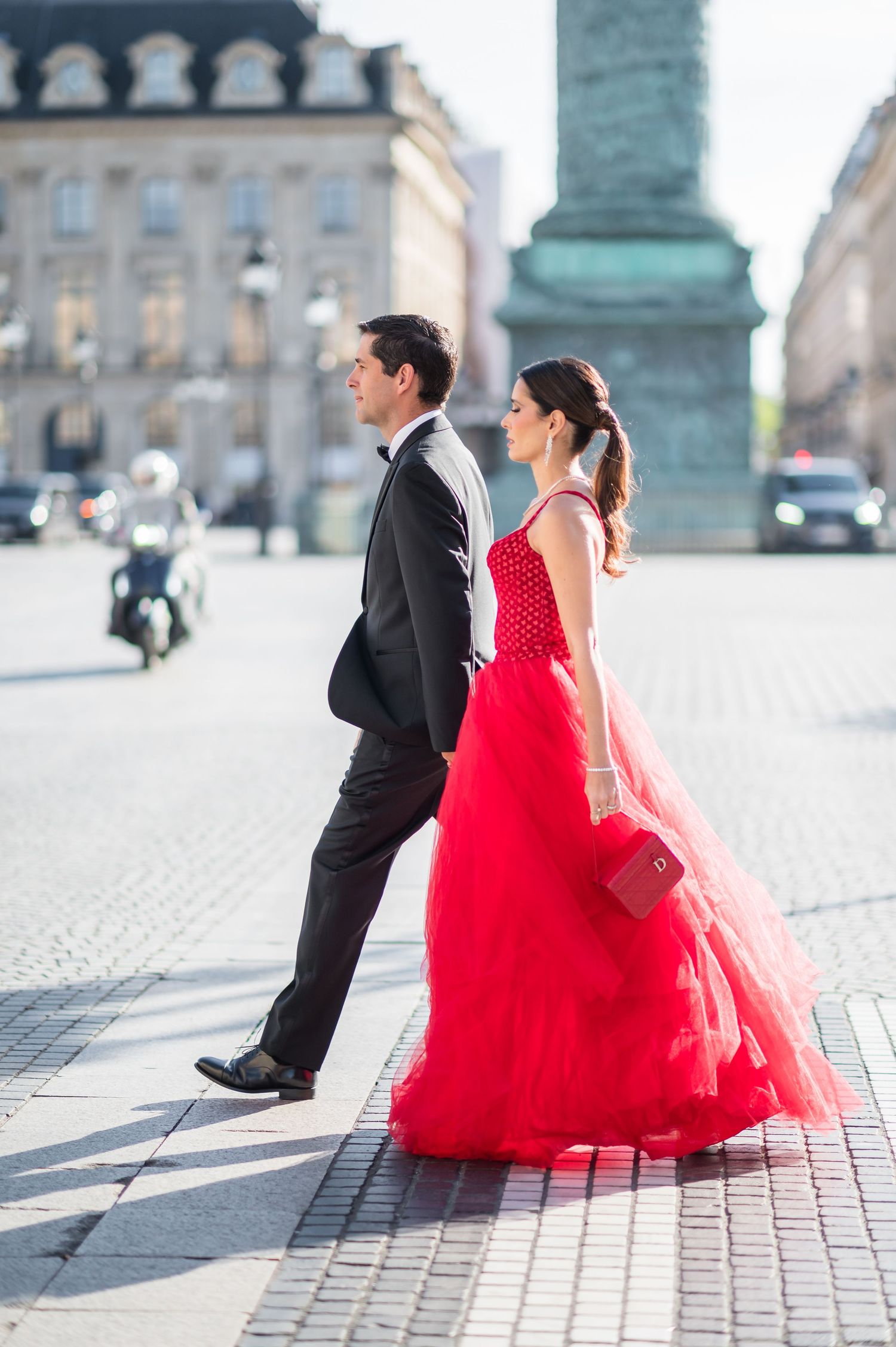 A couple walks across a cobblestone plaza, the woman in a flowing red gown holding a red clutch, the man in a black suit.