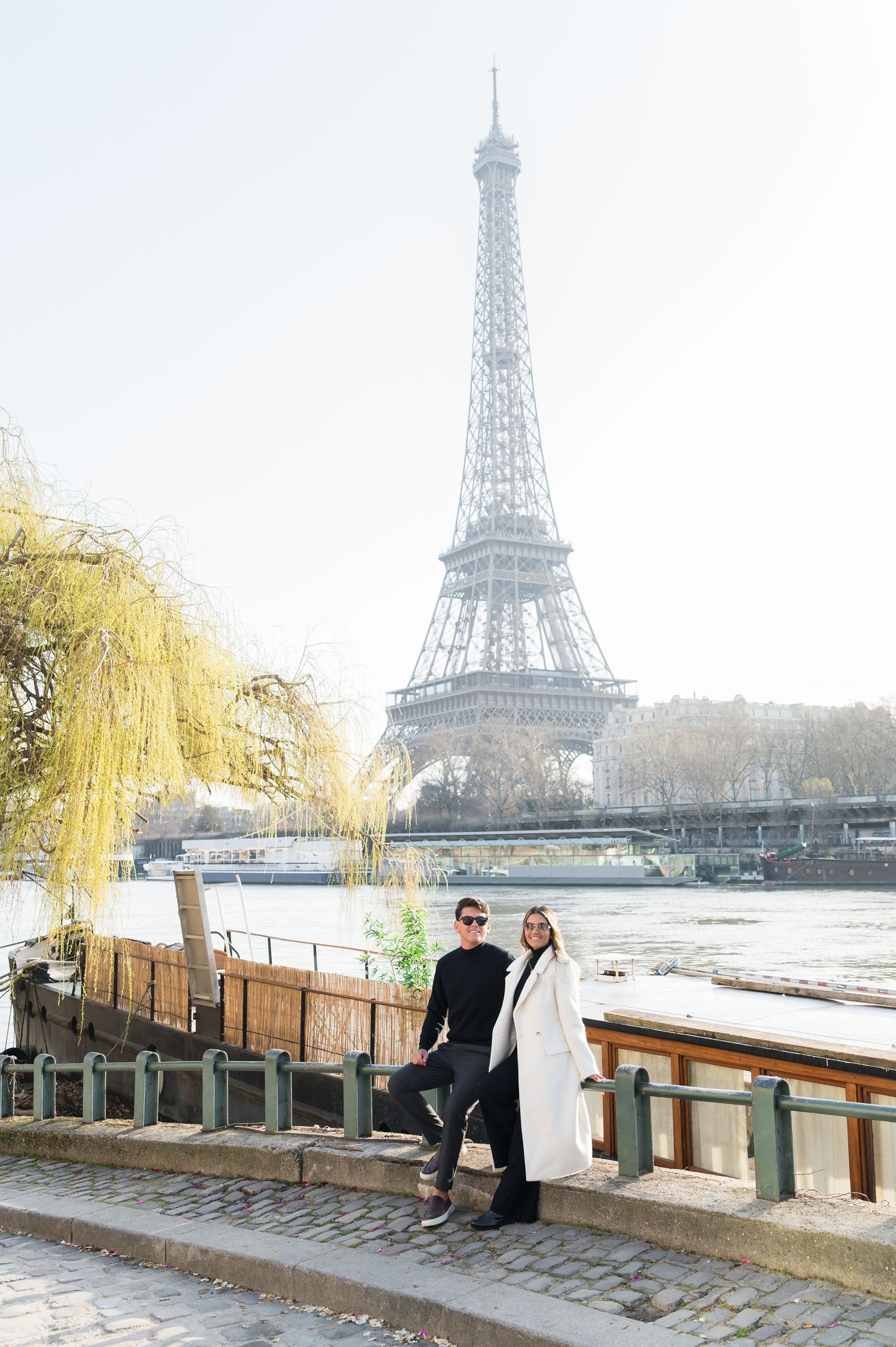 Couple posing by the Seine River with the Eiffel Tower in the background on a bright day in Paris.