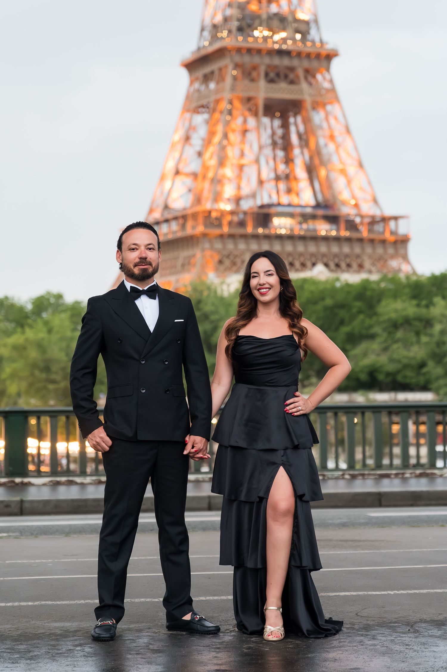 Elegant couple in black formal attire holding hands in front of the illuminated Eiffel Tower in Paris.
