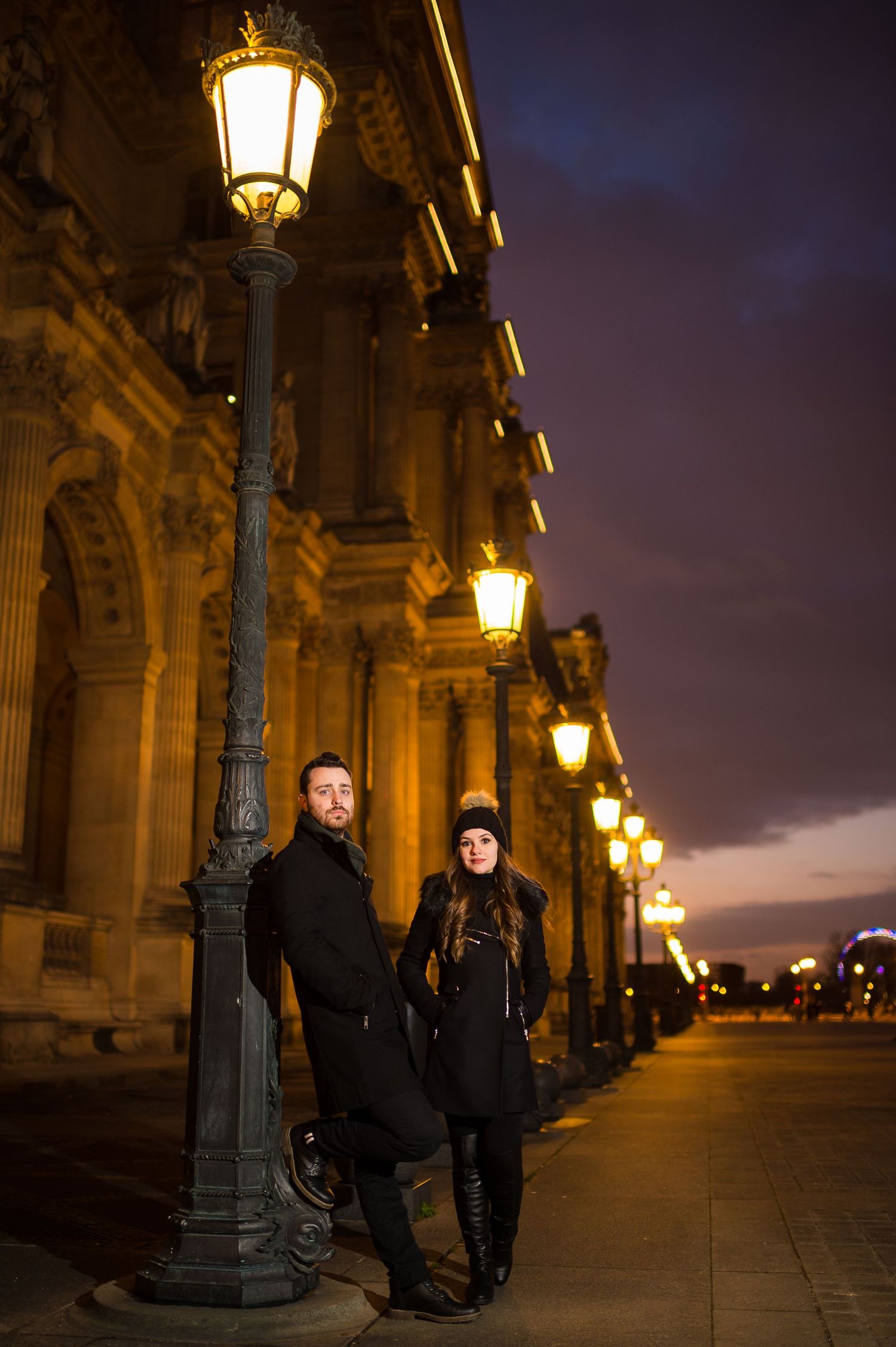 A couple stands beneath illuminated vintage street lamps along an ornate building facade at night in Paris.