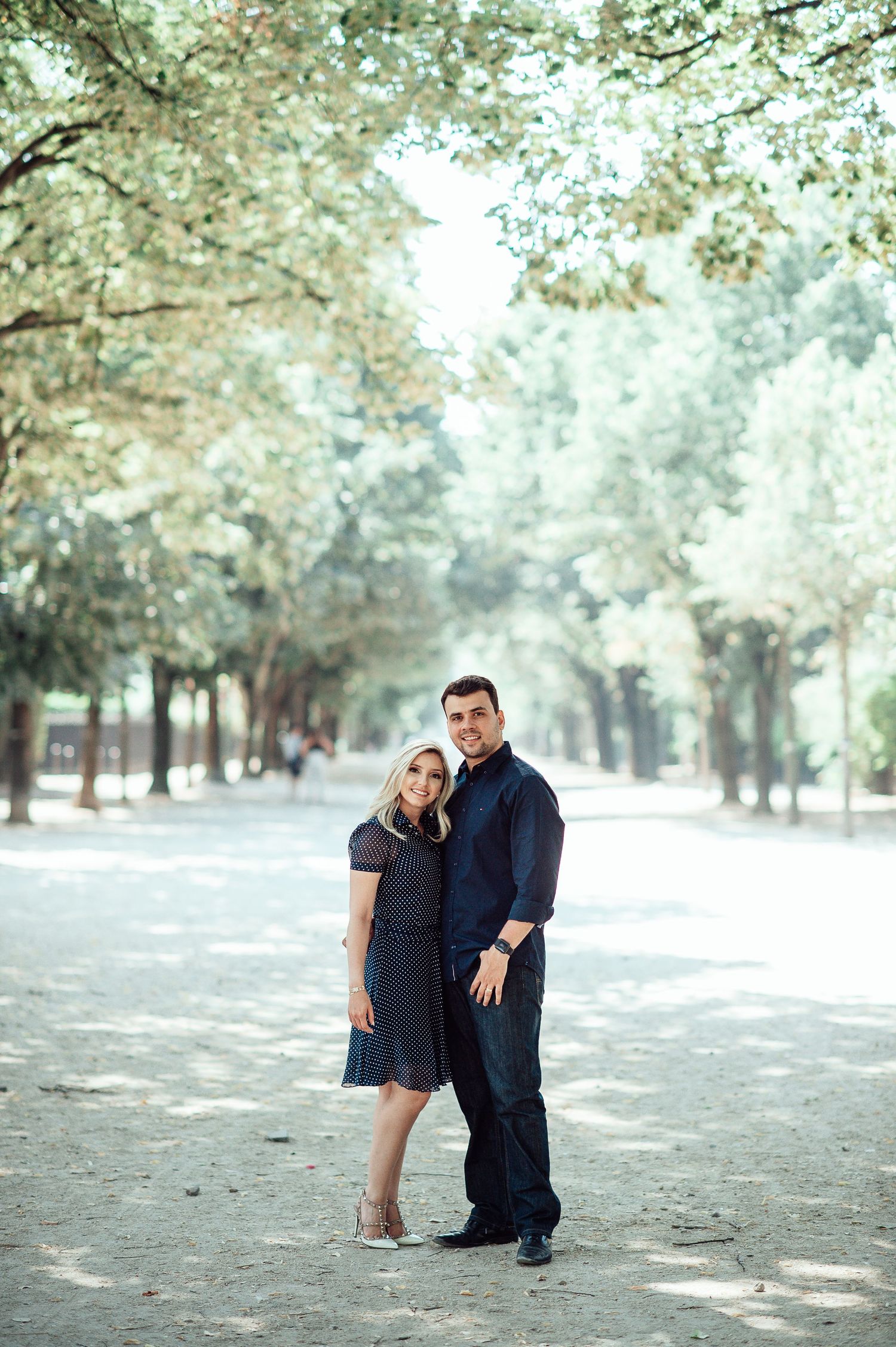 A couple stands lovingly together on a tree-lined path in a park on a bright sunny day.