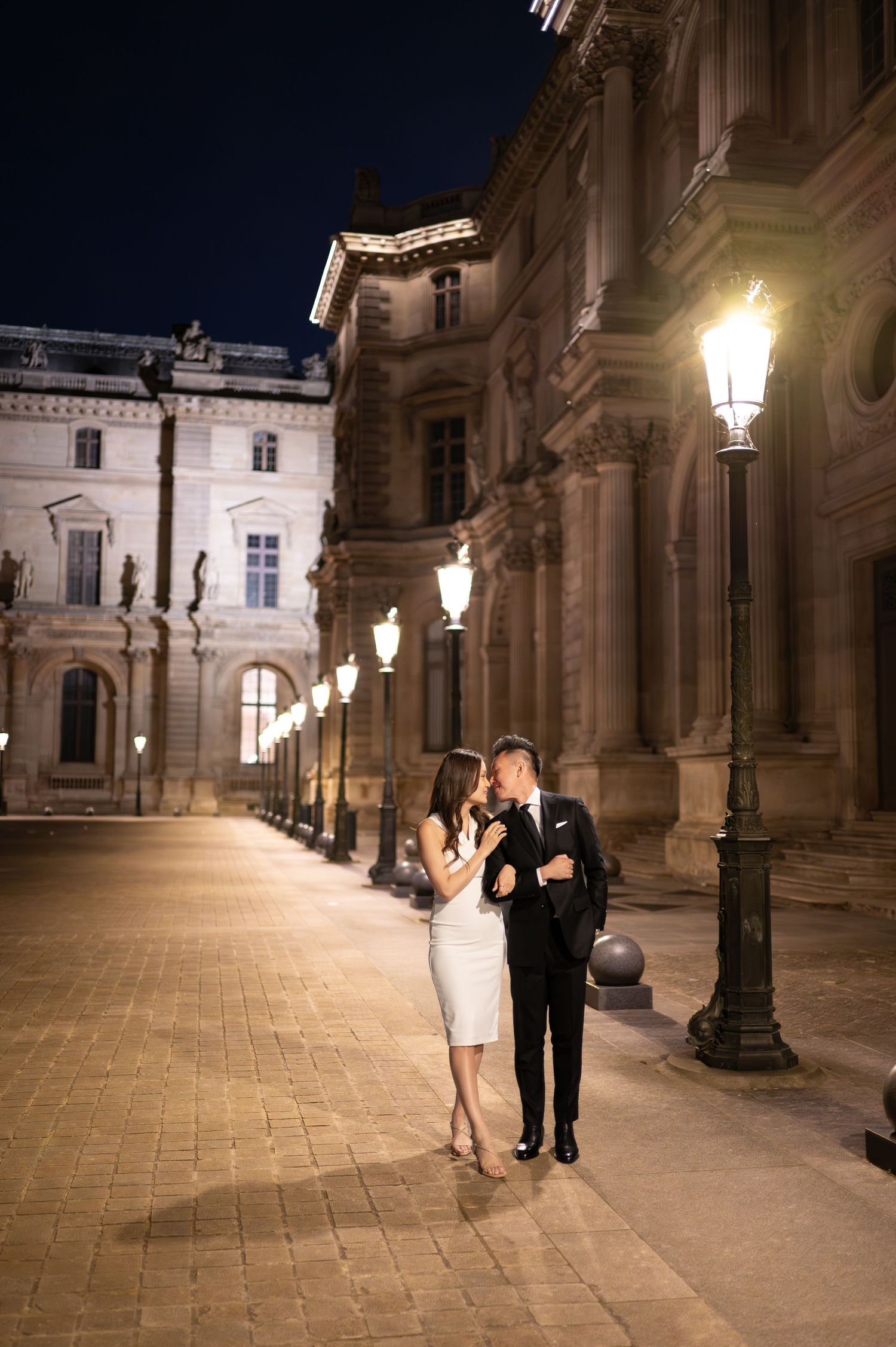 A romantic nighttime scene in Vienna shows a couple walking along illuminated historic architecture with glowing lampposts.
