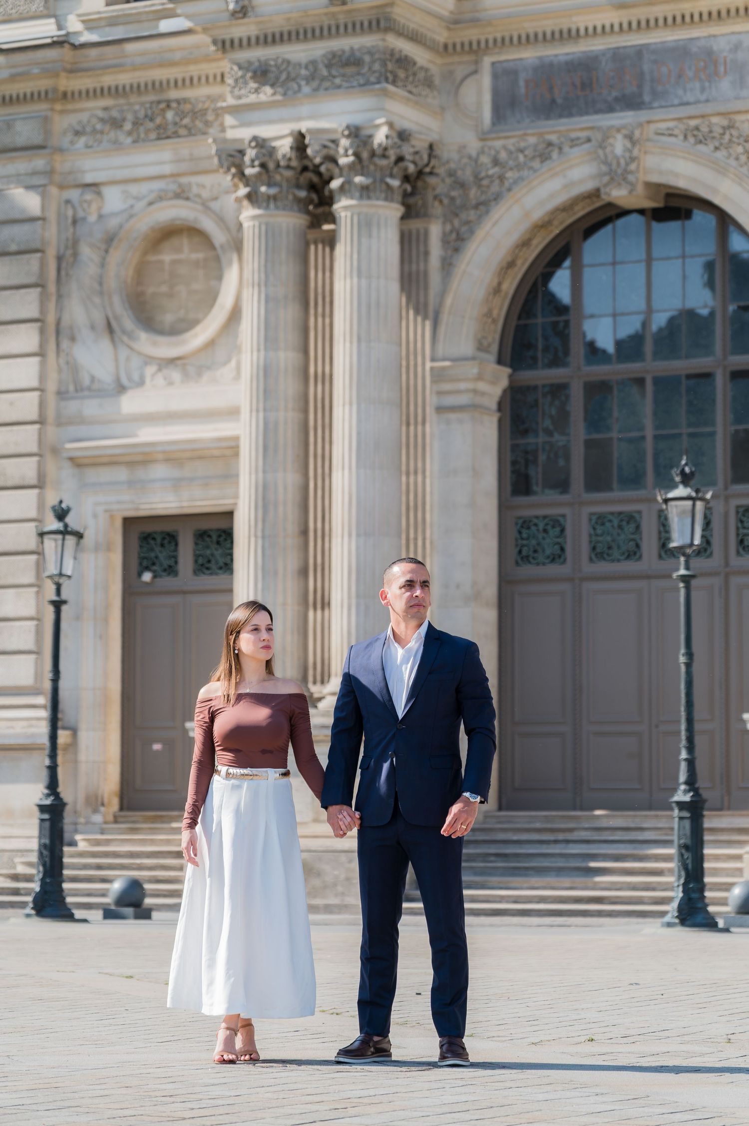 A couple stands playfully in front of an elegant European palace facade with classical architecture and ornate details.