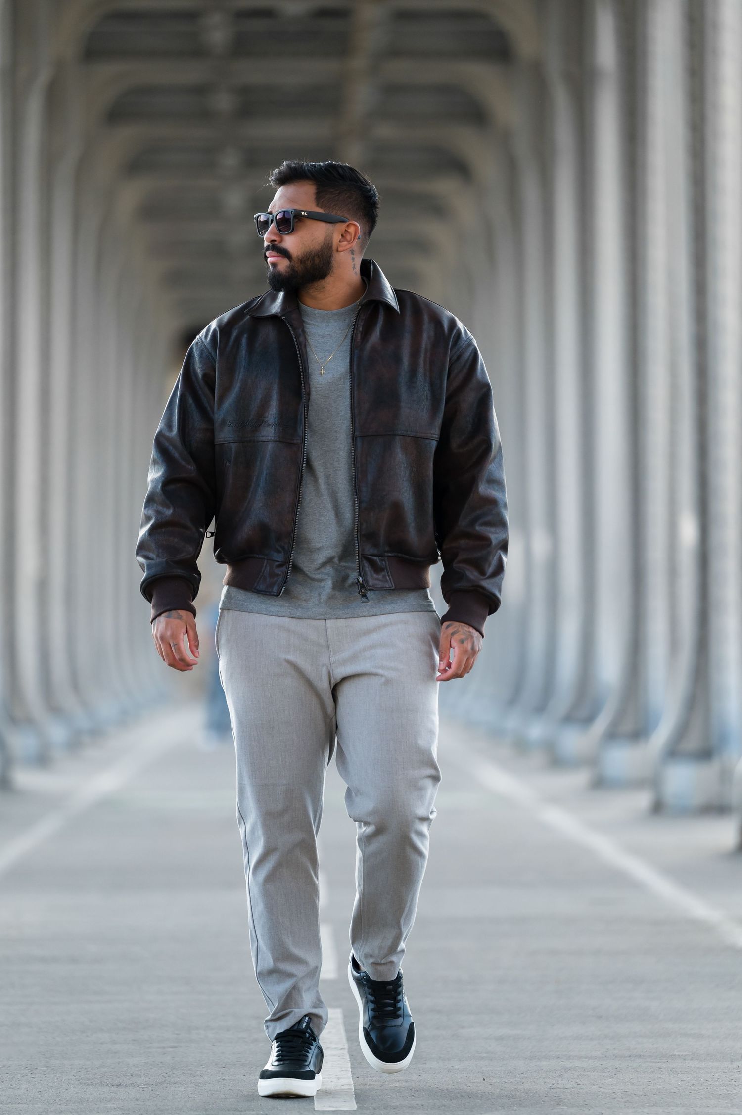 A bearded man in a dark leather jacket, gray joggers, and sunglasses walks confidently through a columned archway.