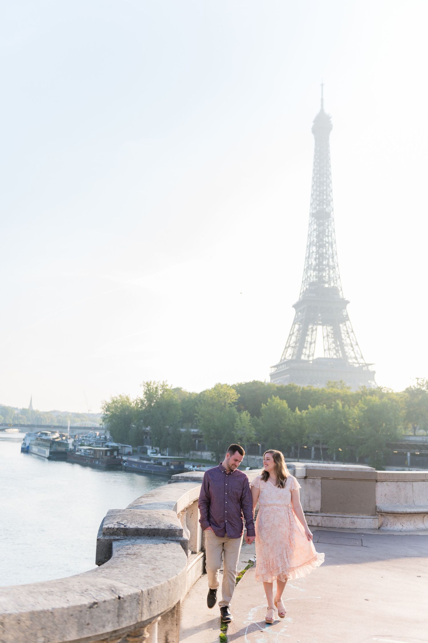 Couple strolling along Seine riverbank with Eiffel Tower and boats visible in background.