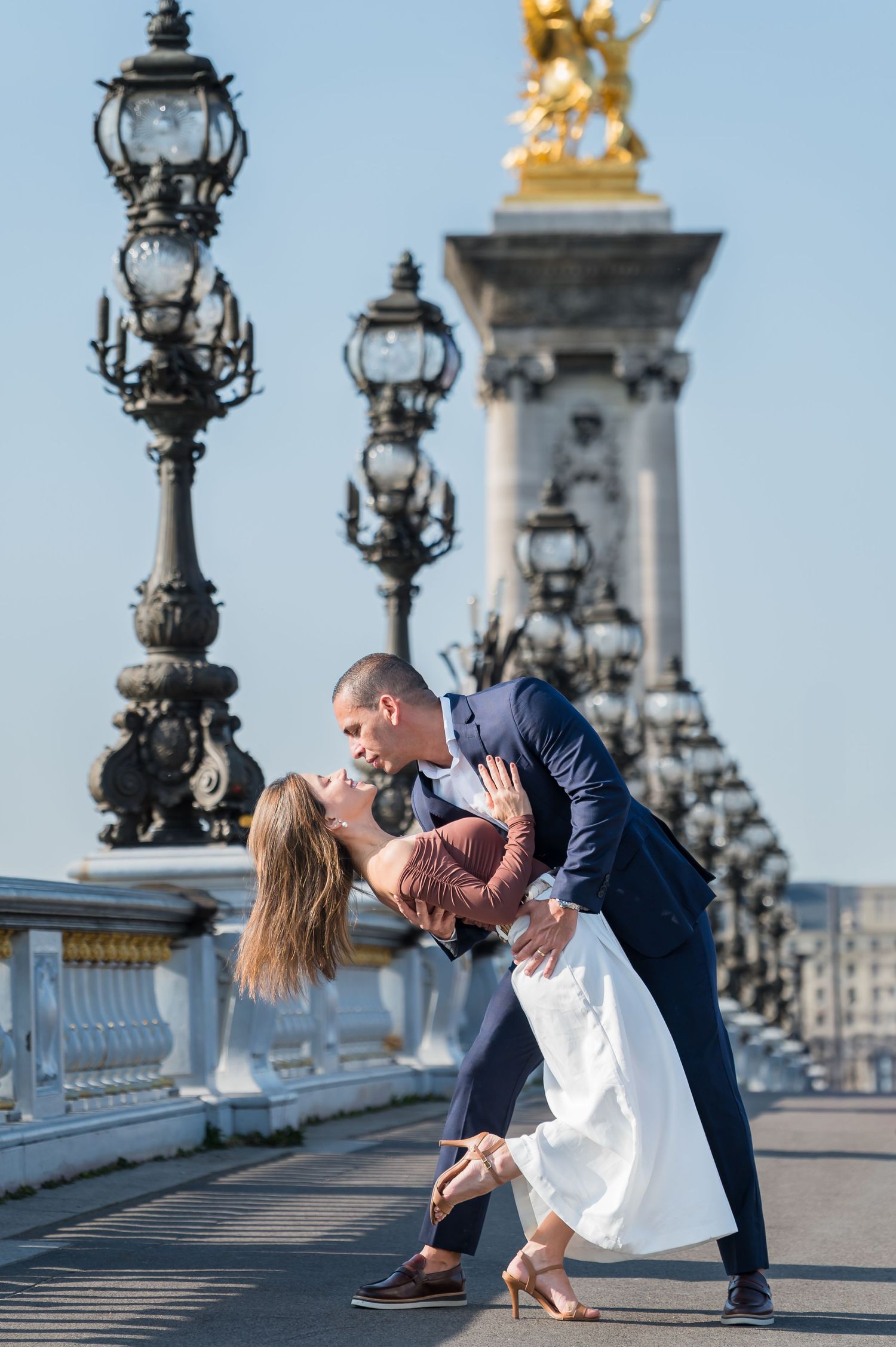 Romantic couple dancing on scenic bridge with vintage lampposts and golden statue in background.