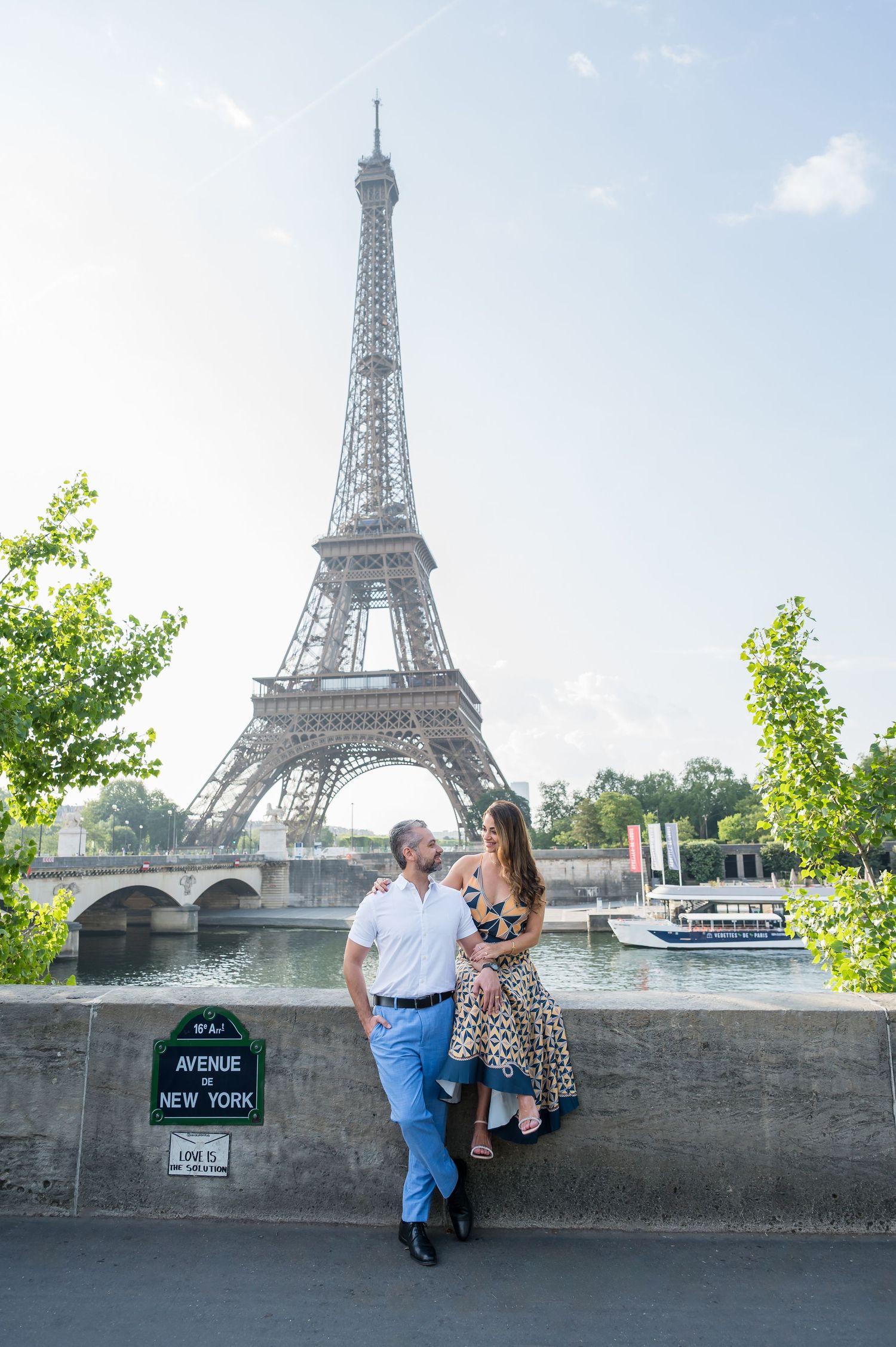 A couple poses along the Seine River with the iconic Eiffel Tower and a tour boat in the background in Paris.