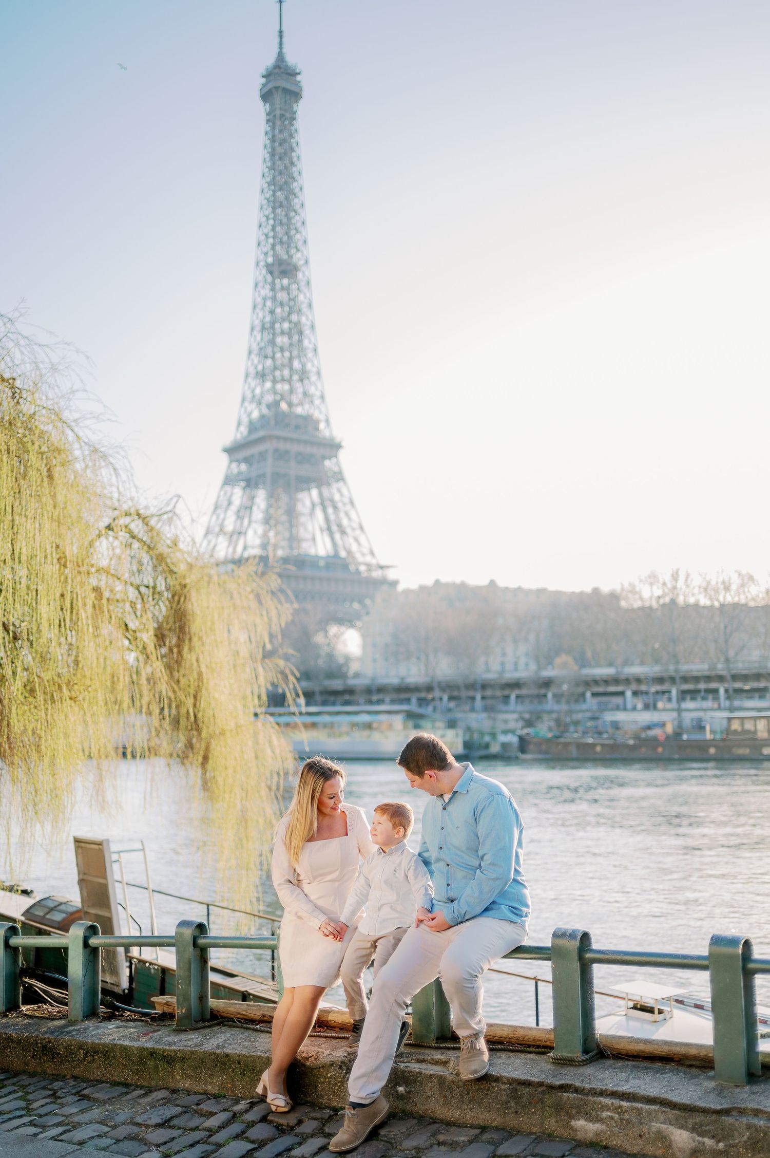 Family sits together on riverside railing with Eiffel Tower and willow tree in view.