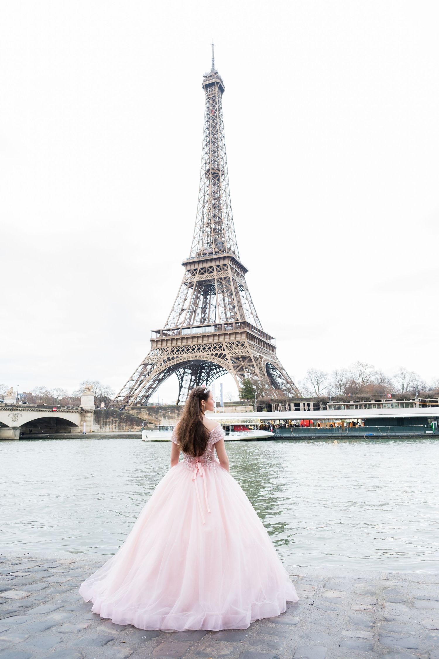 A person in a flowing pink ball gown stands by the Seine River looking at the Eiffel Tower in Paris.