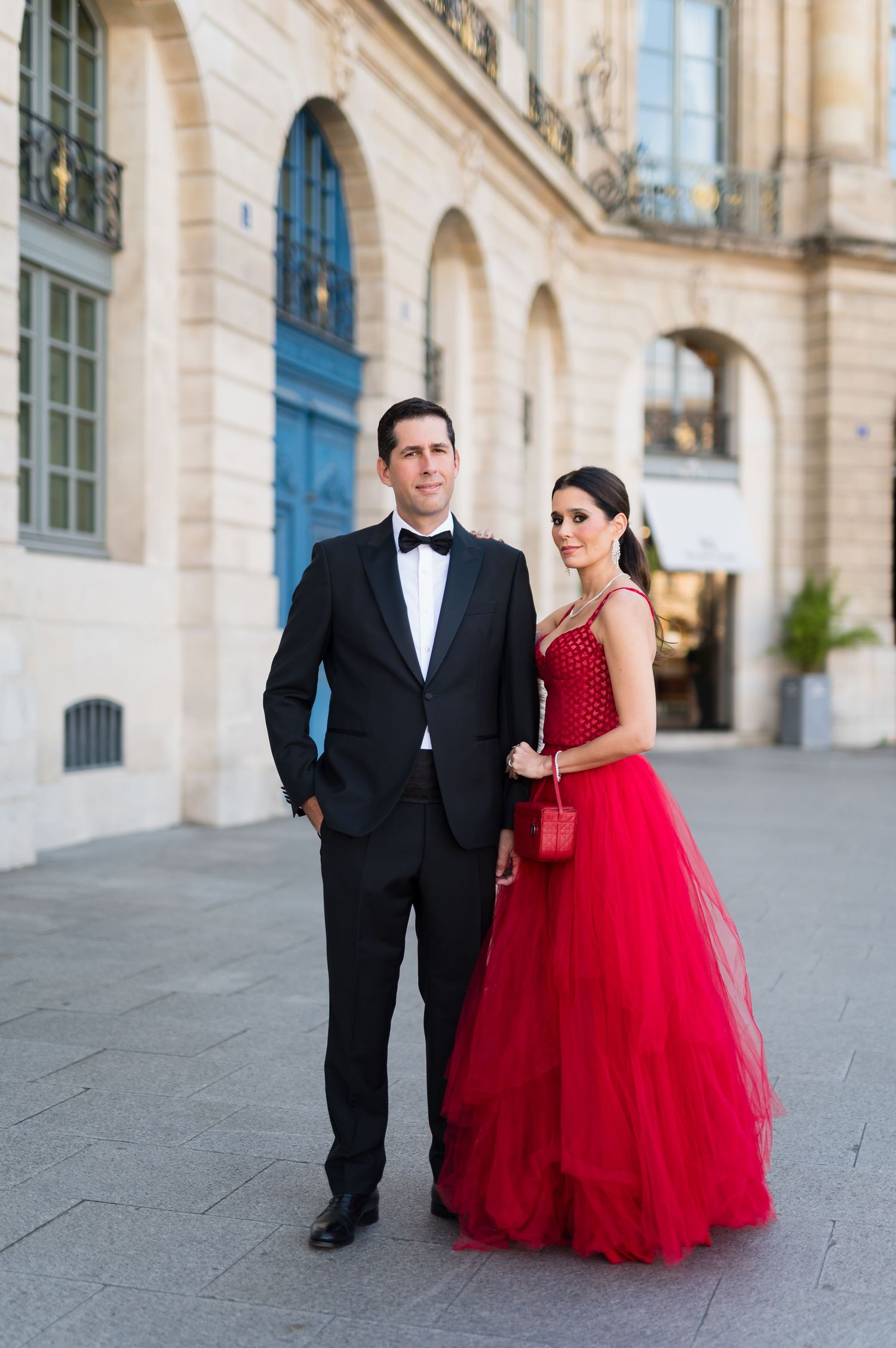 Elegant couple in formal attire, man in tuxedo and woman in red ball gown, posing outside a classic European building.