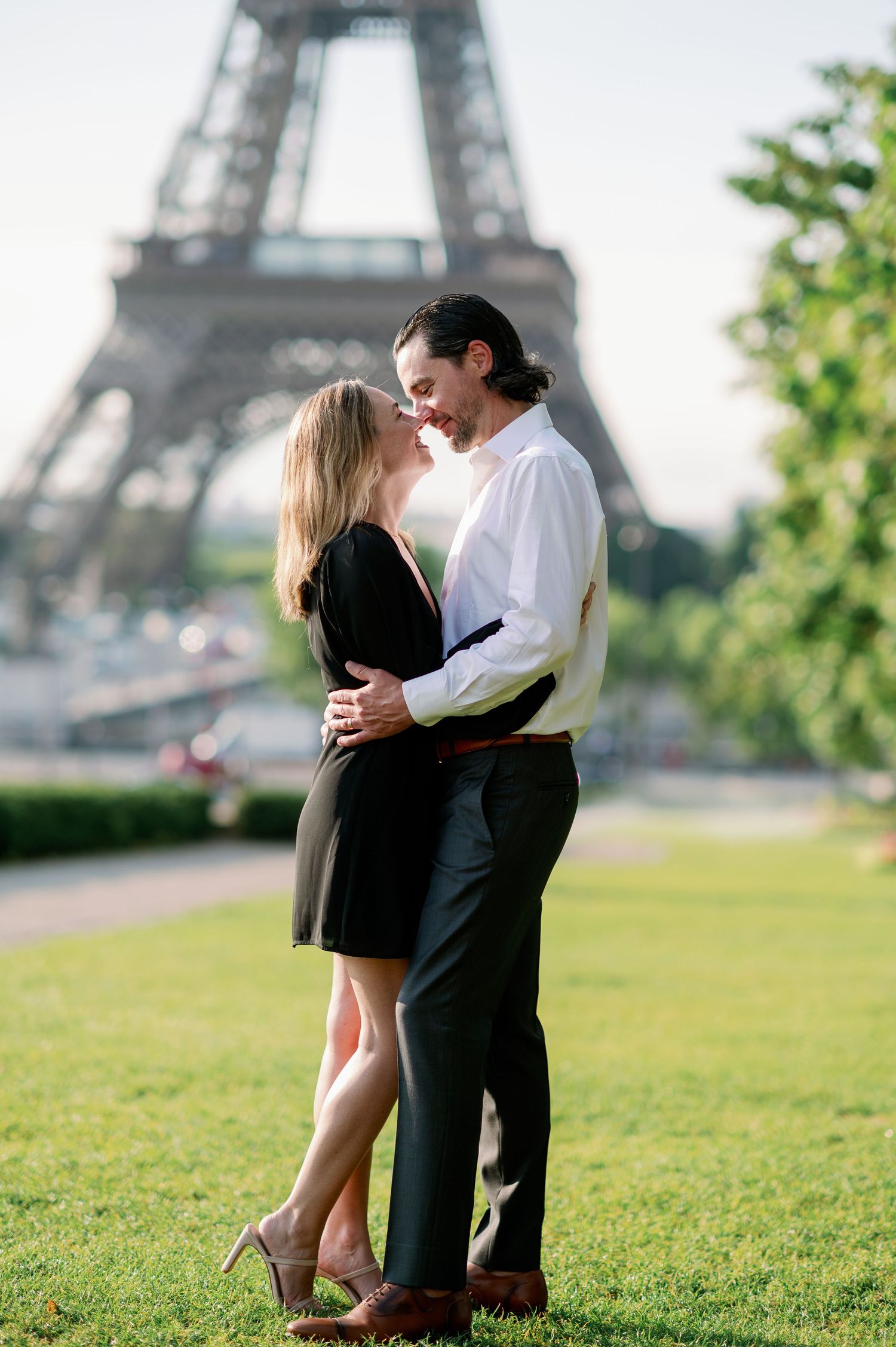 Romantic couple embracing in front of the Eiffel Tower in Paris, woman in black dress, man in white shirt.