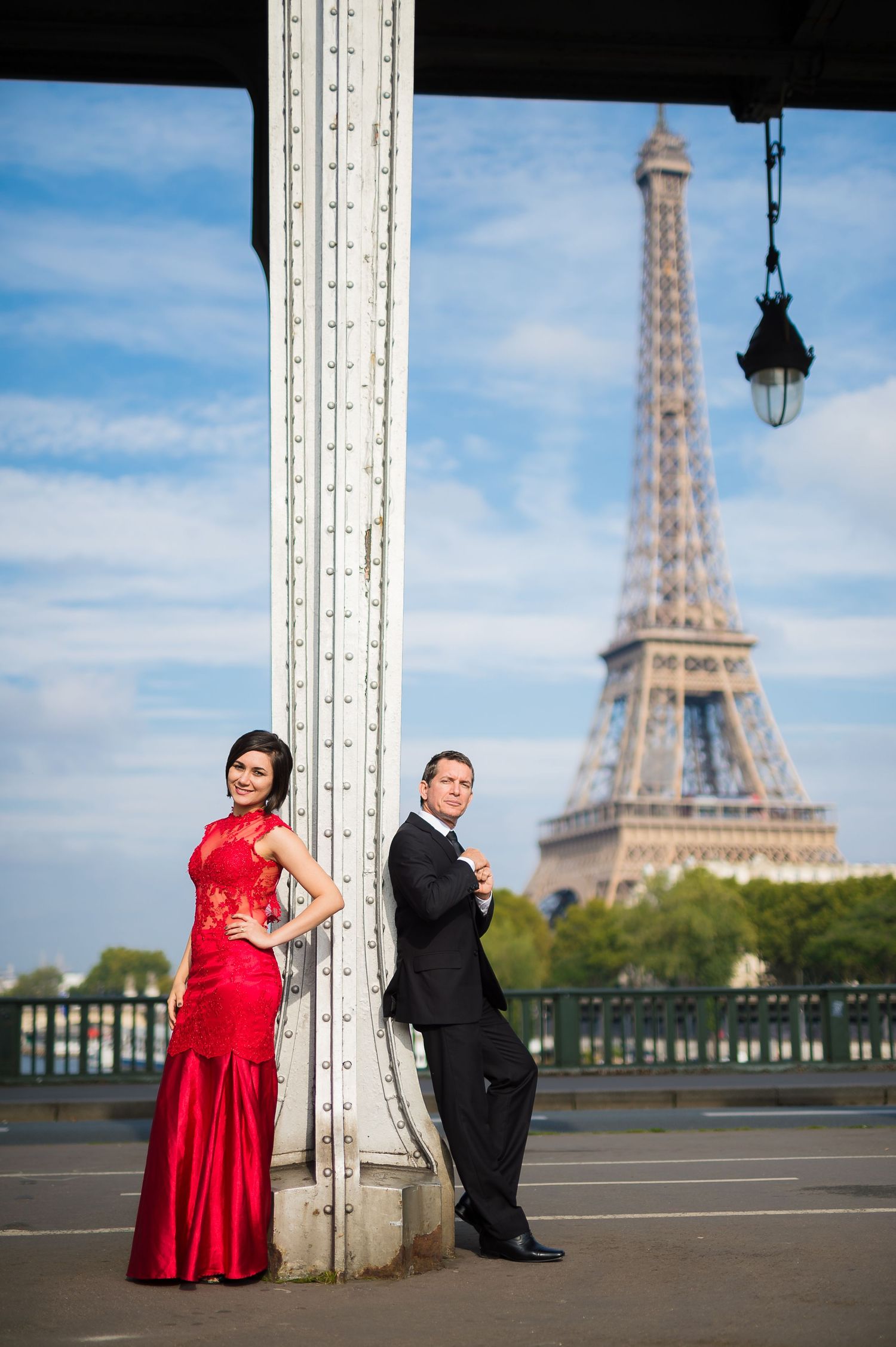Couple in formal wear posing by Eiffel Tower in Paris, woman in red gown, man in black suit.