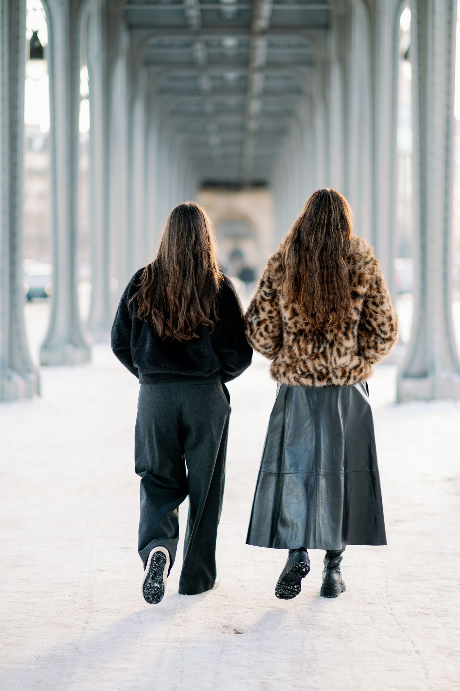Two people walking down a columned corridor wearing winter coats, one in black and one in leopard print.