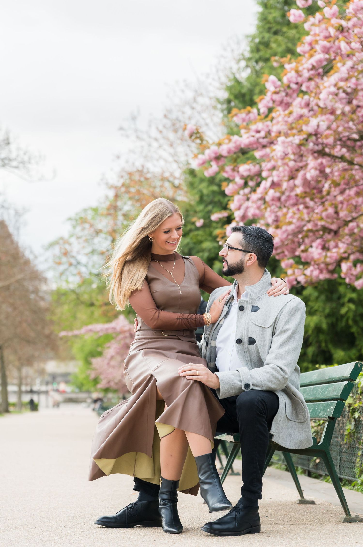 Couple sitting on a park bench surrounded by pink cherry blossoms, sharing a romantic moment in spring.