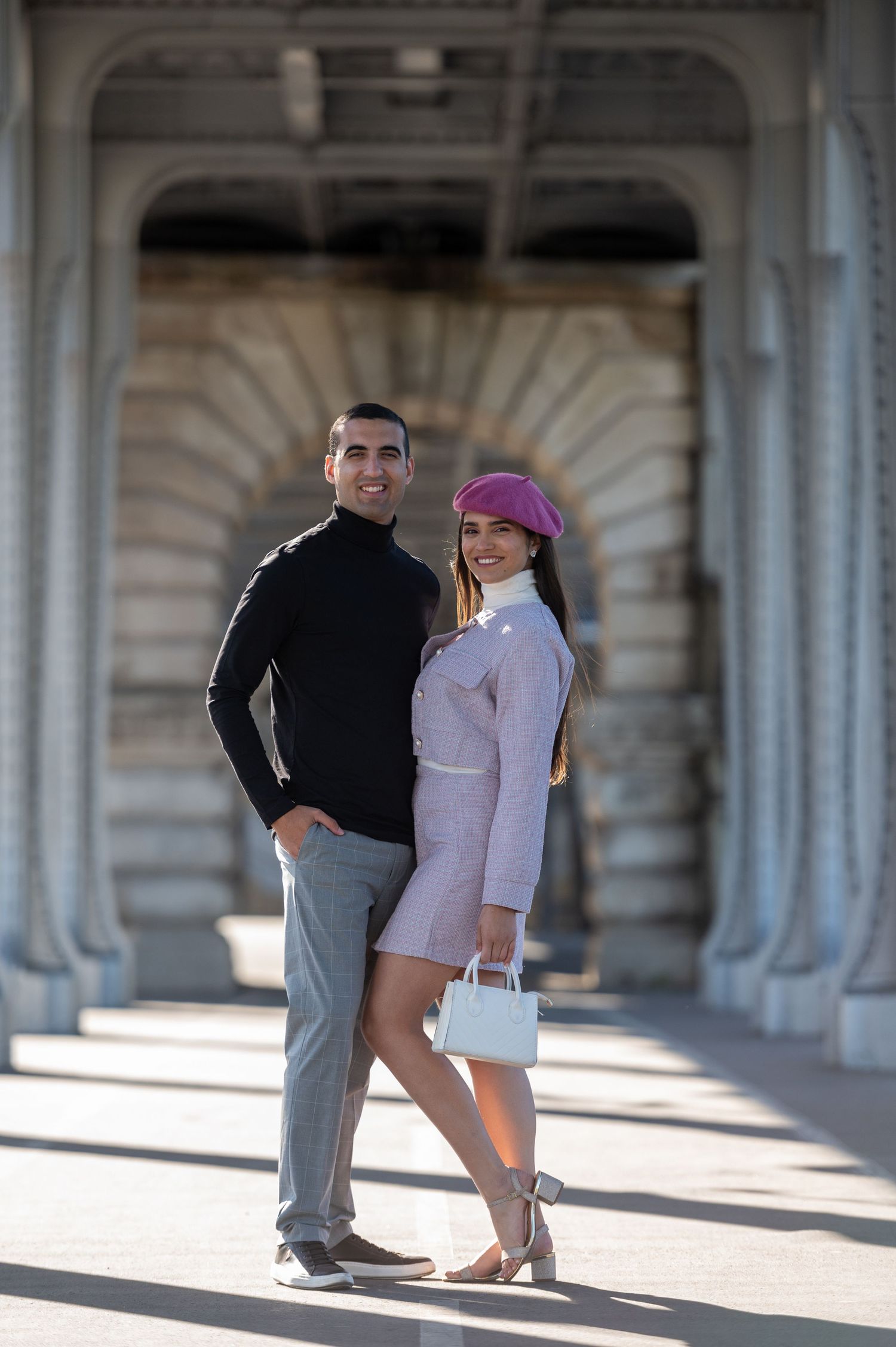 A fashionable couple posing elegantly under an ornate archway in Paris, with the woman wearing a purple beret and white bag.