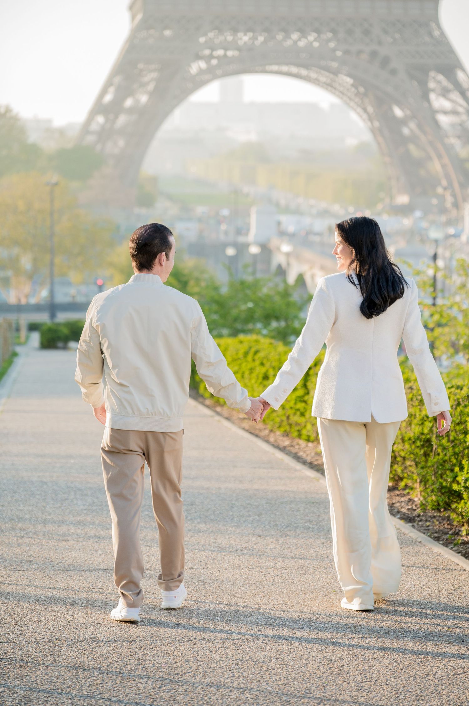 Romantic couple in matching white outfits walking together along a scenic path near the Eiffel Tower in Paris.