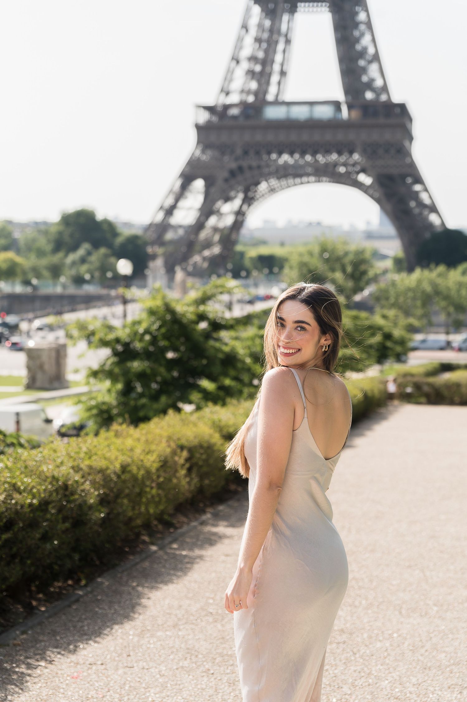 A smiling woman in a white slip dress poses on a path near the Eiffel Tower in Paris, France.