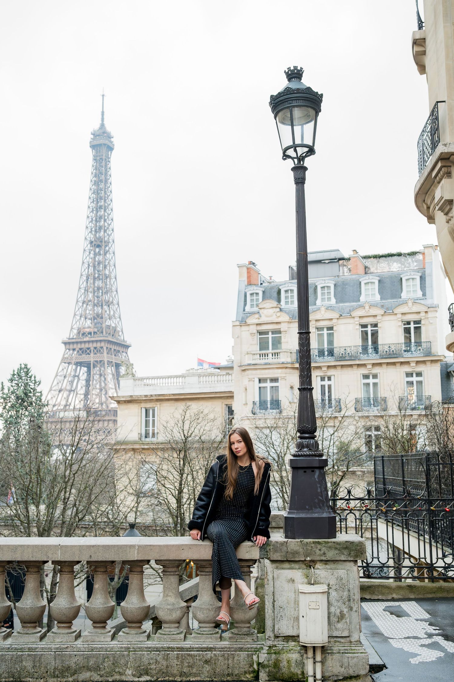 Person sitting on stone balustrade near vintage lamp post with Eiffel Tower and Parisian buildings.