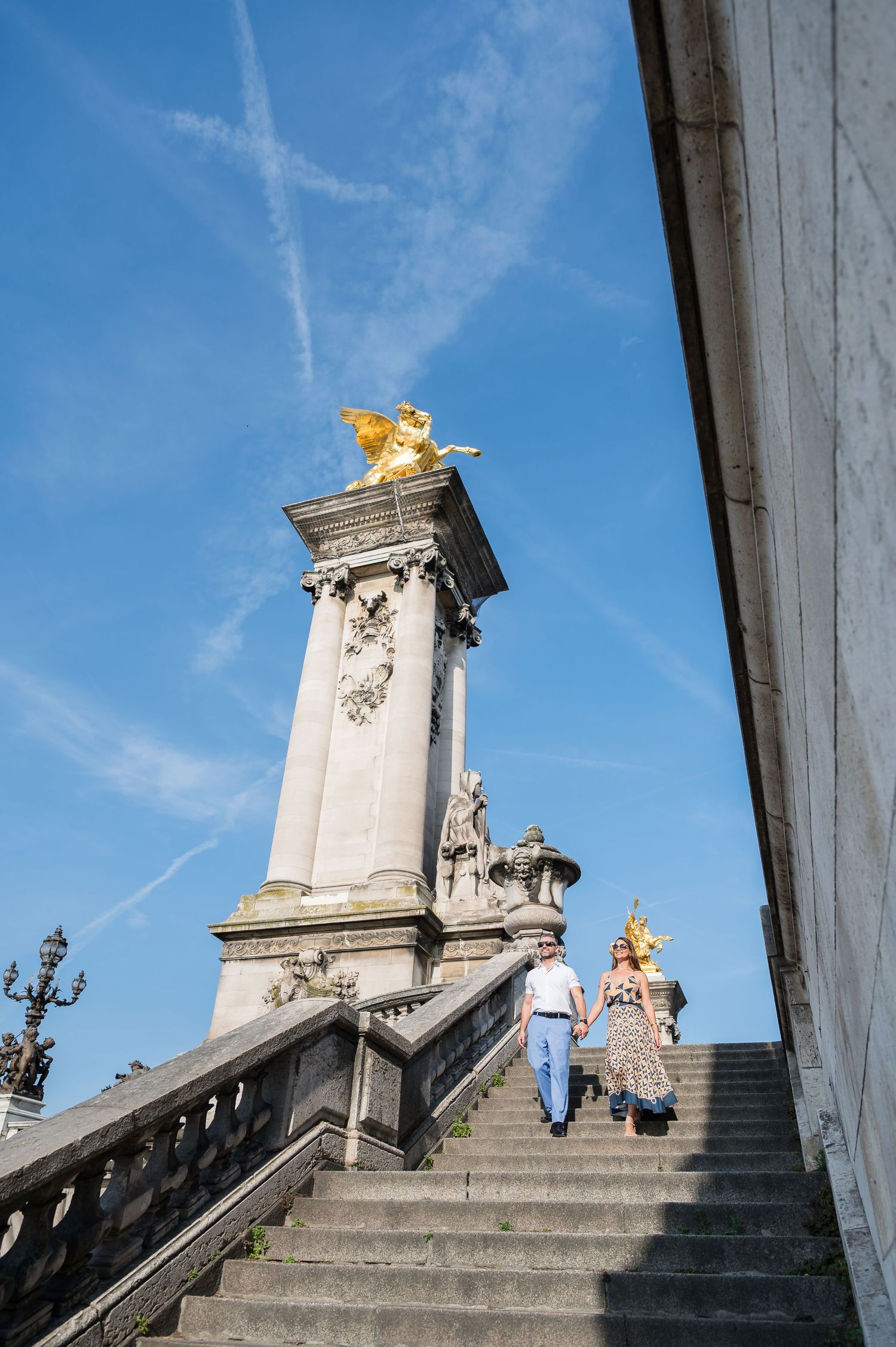 People climbing white marble steps flanked by statues and gold accents on what appears to be a palace entrance on a sunny day.