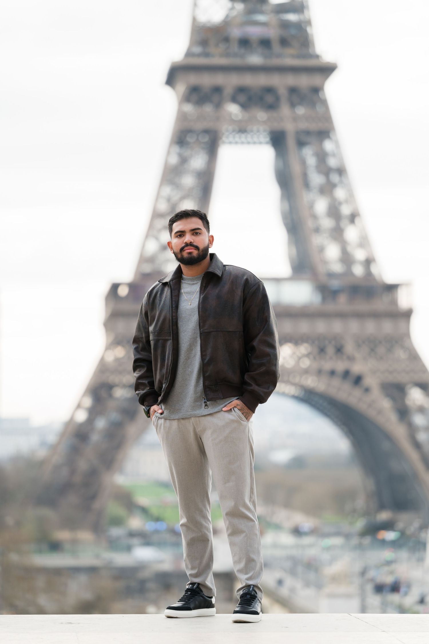 A bearded man in a brown leather jacket poses confidently in front of the Eiffel Tower in Paris, France.