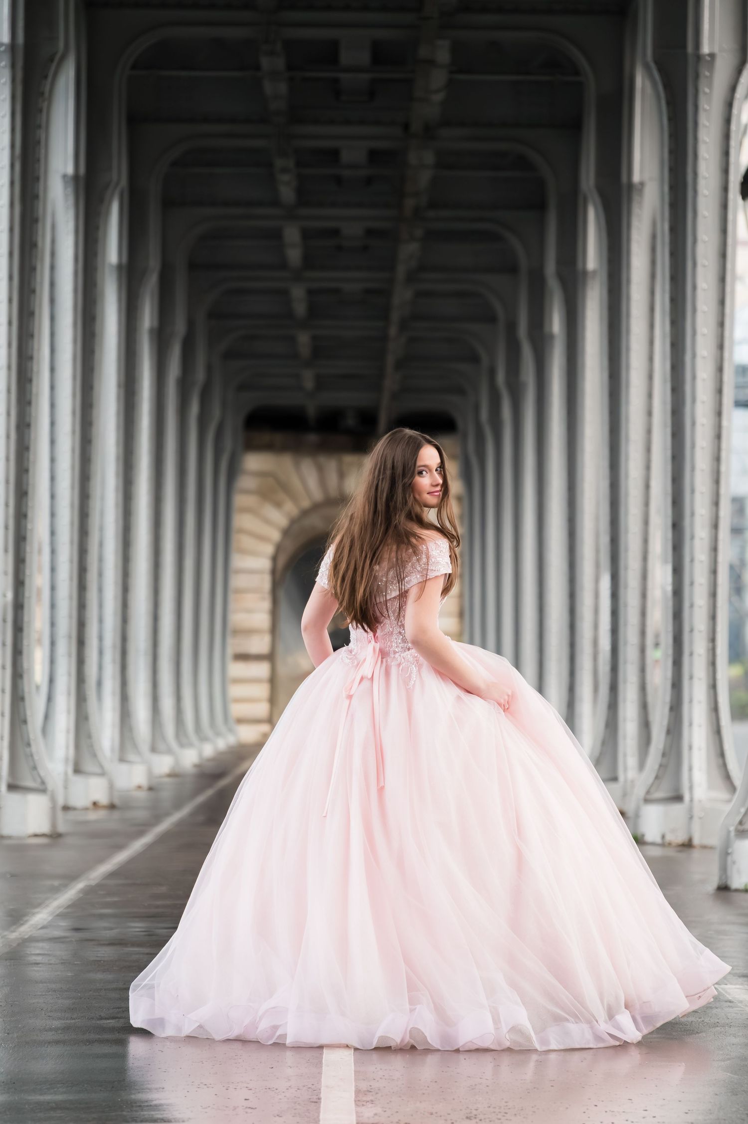 A person in a flowing pink tulle gown poses gracefully under an ornate covered walkway with arched columns.