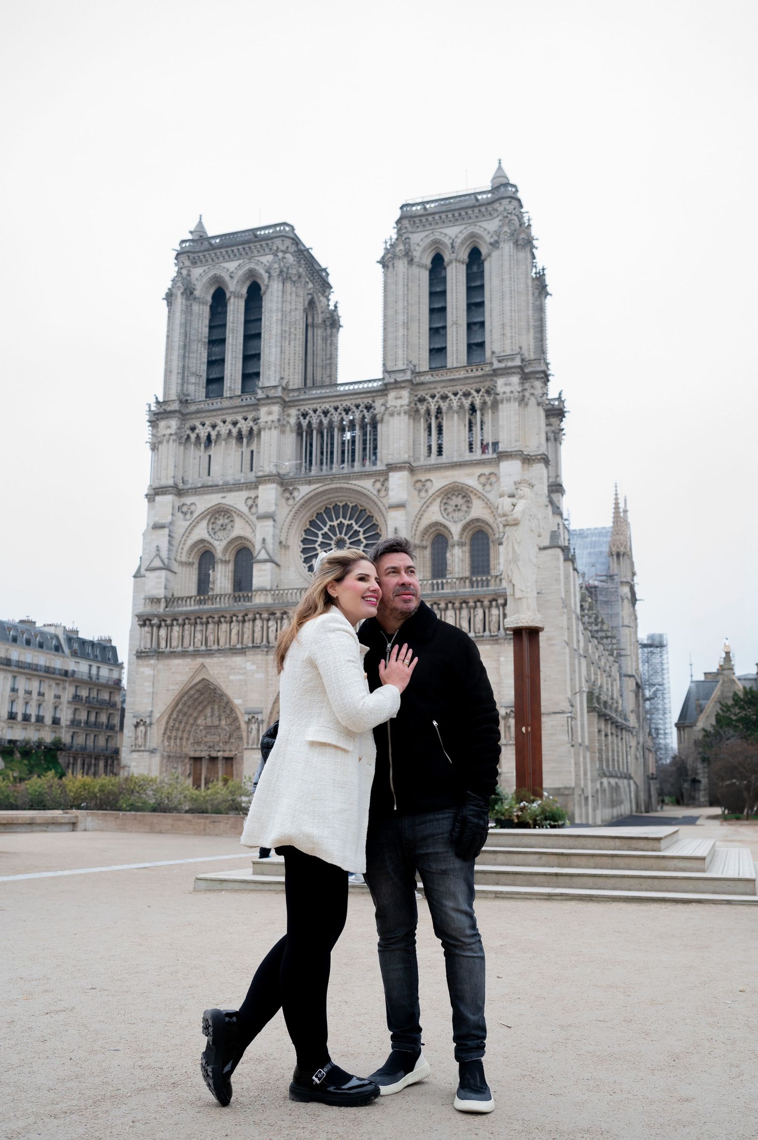 A couple poses for a photo in front of the iconic Notre Dame Cathedral in Paris on a cloudy day.