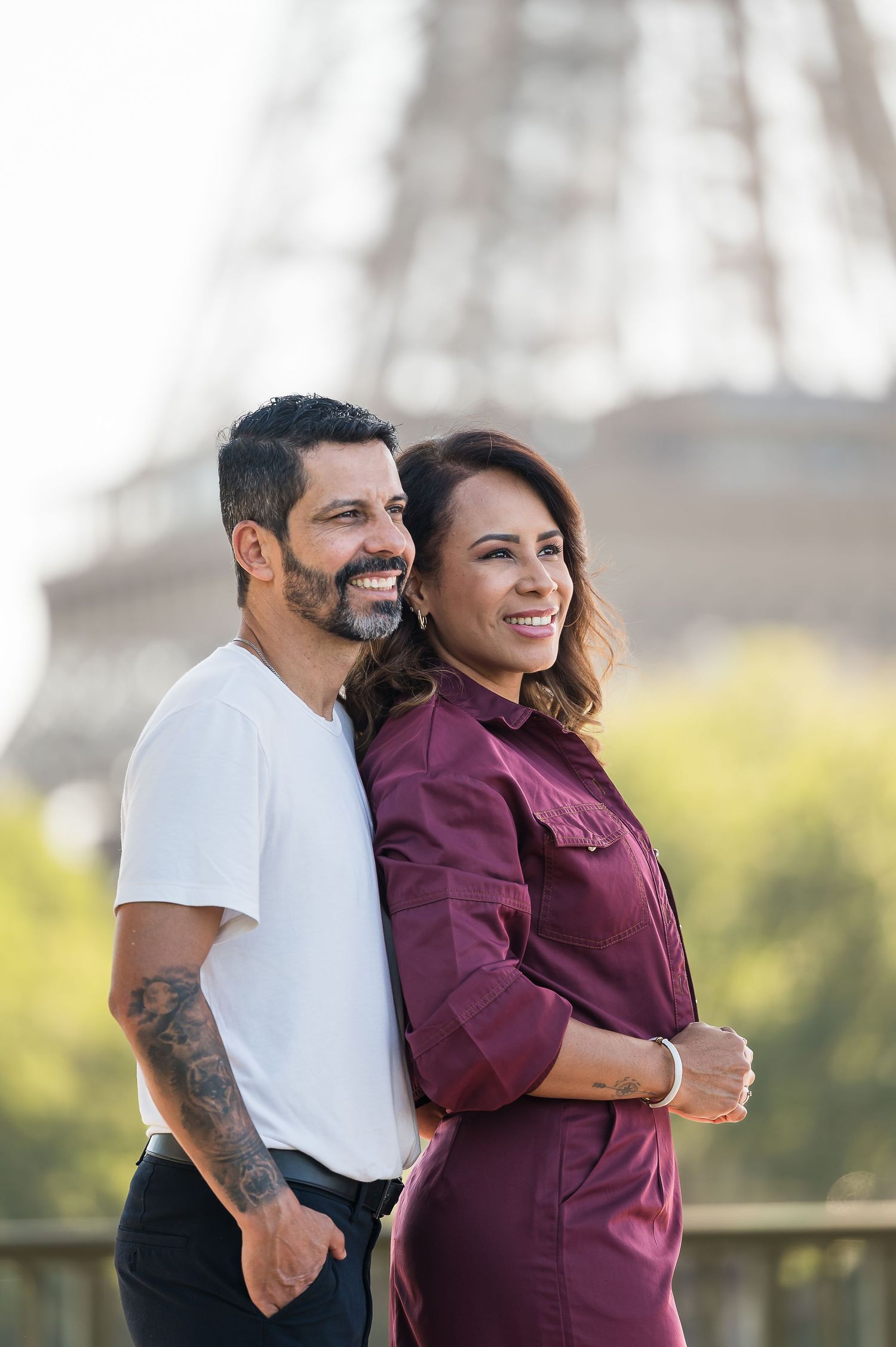 Smiling couple posing together in front of the Eiffel Tower in Paris, man with tattoo in white tee, woman in burgundy jumpsuit.