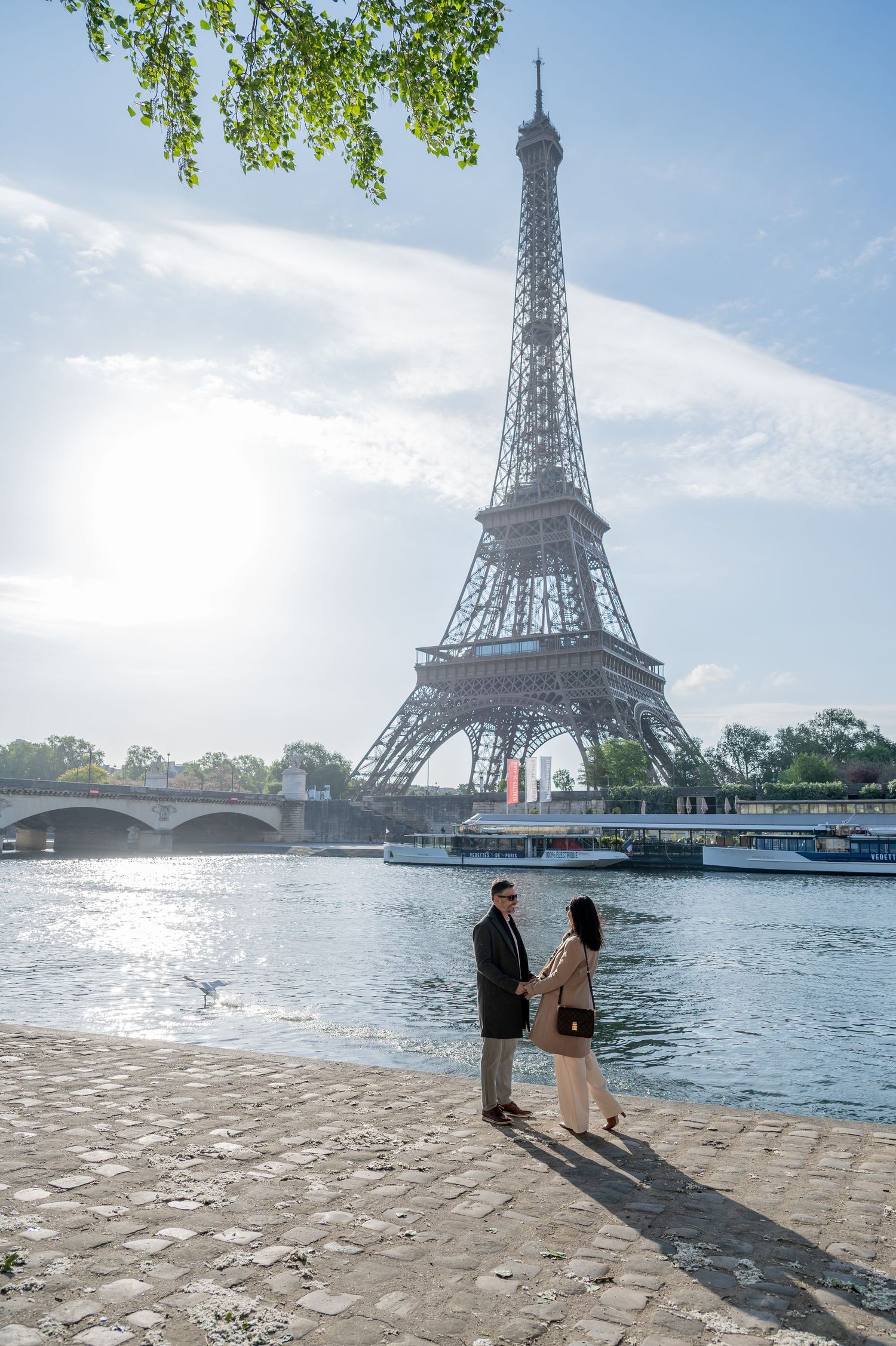 Romantic couple admiring Eiffel Tower view along Seine River on cobblestone path in Paris.