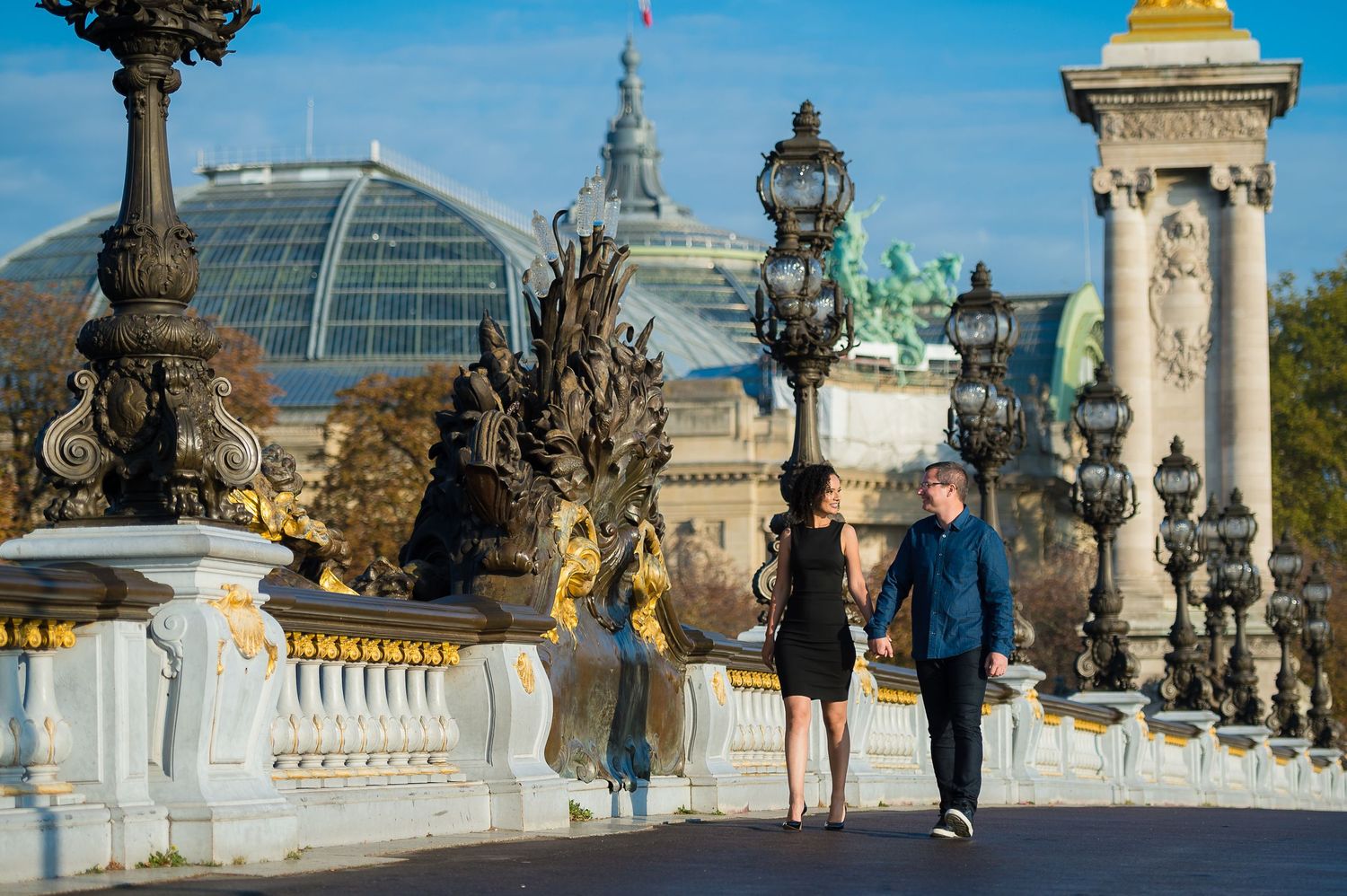 Two people stroll across the ornate Alexandre III Bridge in Paris with Grand Palais dome visible in background.