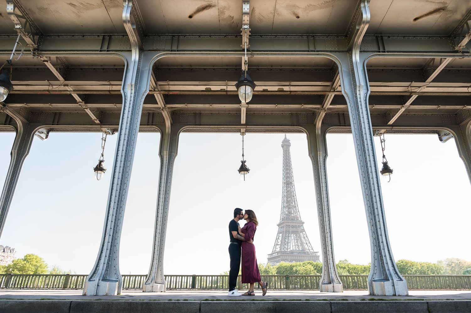 Couple kissing under Bir-Hakeim Bridge with Eiffel Tower in background, Paris romantic engagement photo.