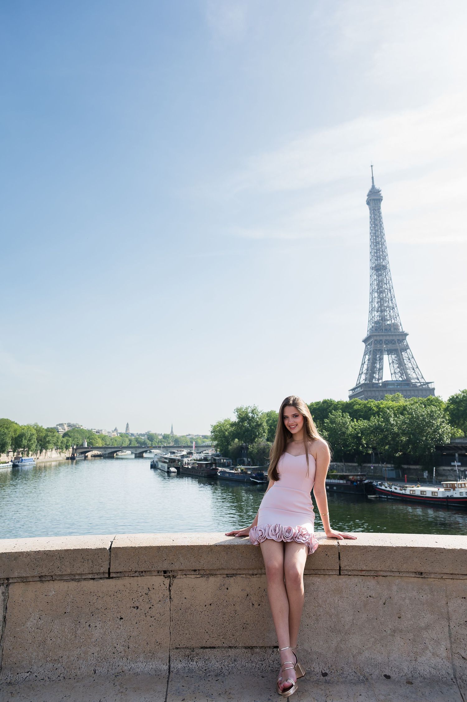 Young woman in pink dress sitting on a stone wall by the Seine River with the Eiffel Tower in Paris, France.