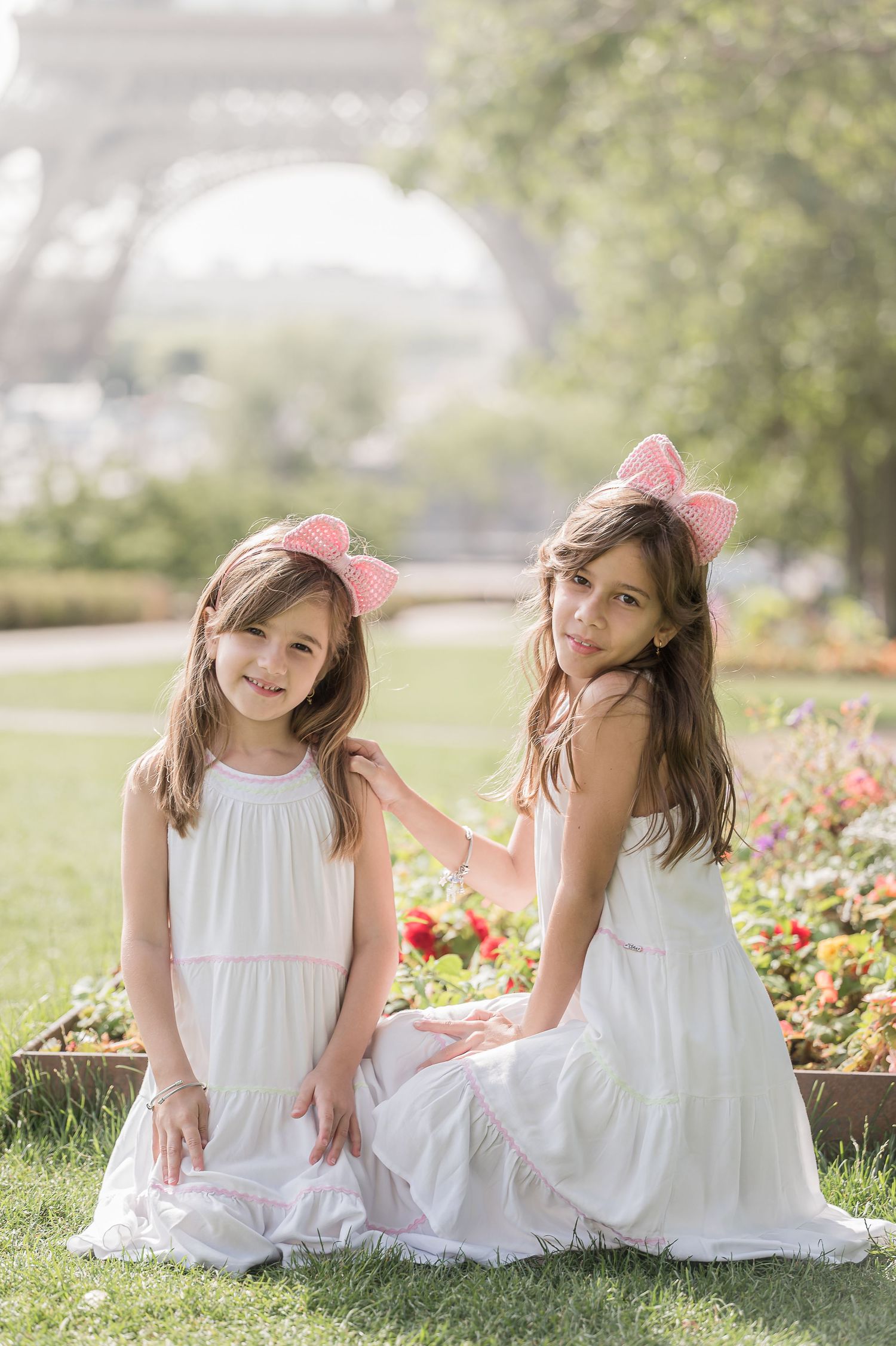 Two young girls in white dresses with pink bow headbands pose in a park near the Eiffel Tower in Paris.