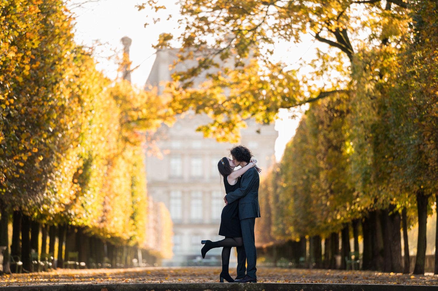 Couple sharing a romantic kiss in a golden autumn park alley with yellow trees and a classical building backdrop.