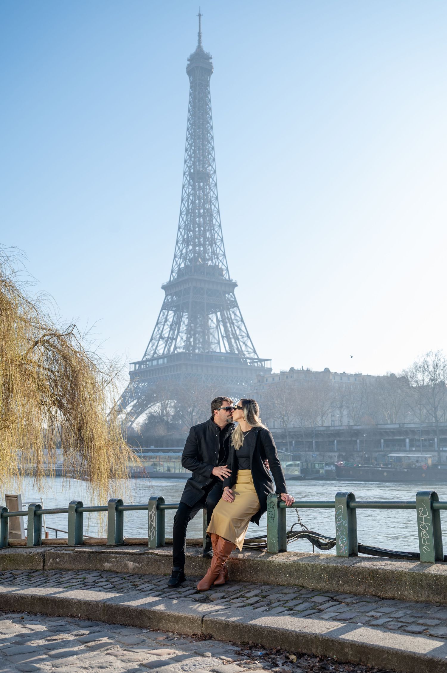 Romantic couple pose on riverside wall with Eiffel Tower in background.