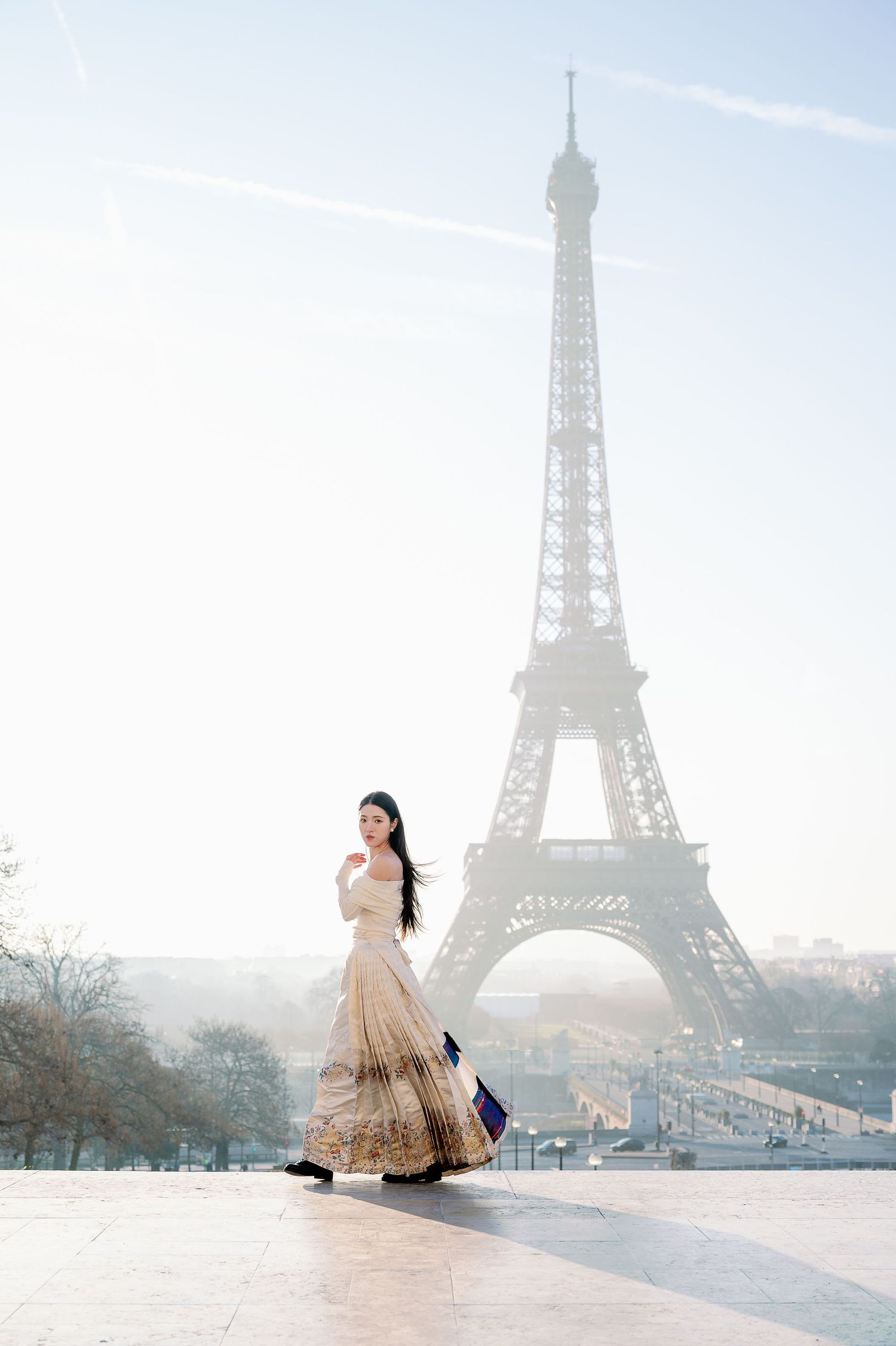 A dreamy portrait of a woman in a beige gown with the Eiffel Tower emerging through morning fog.
