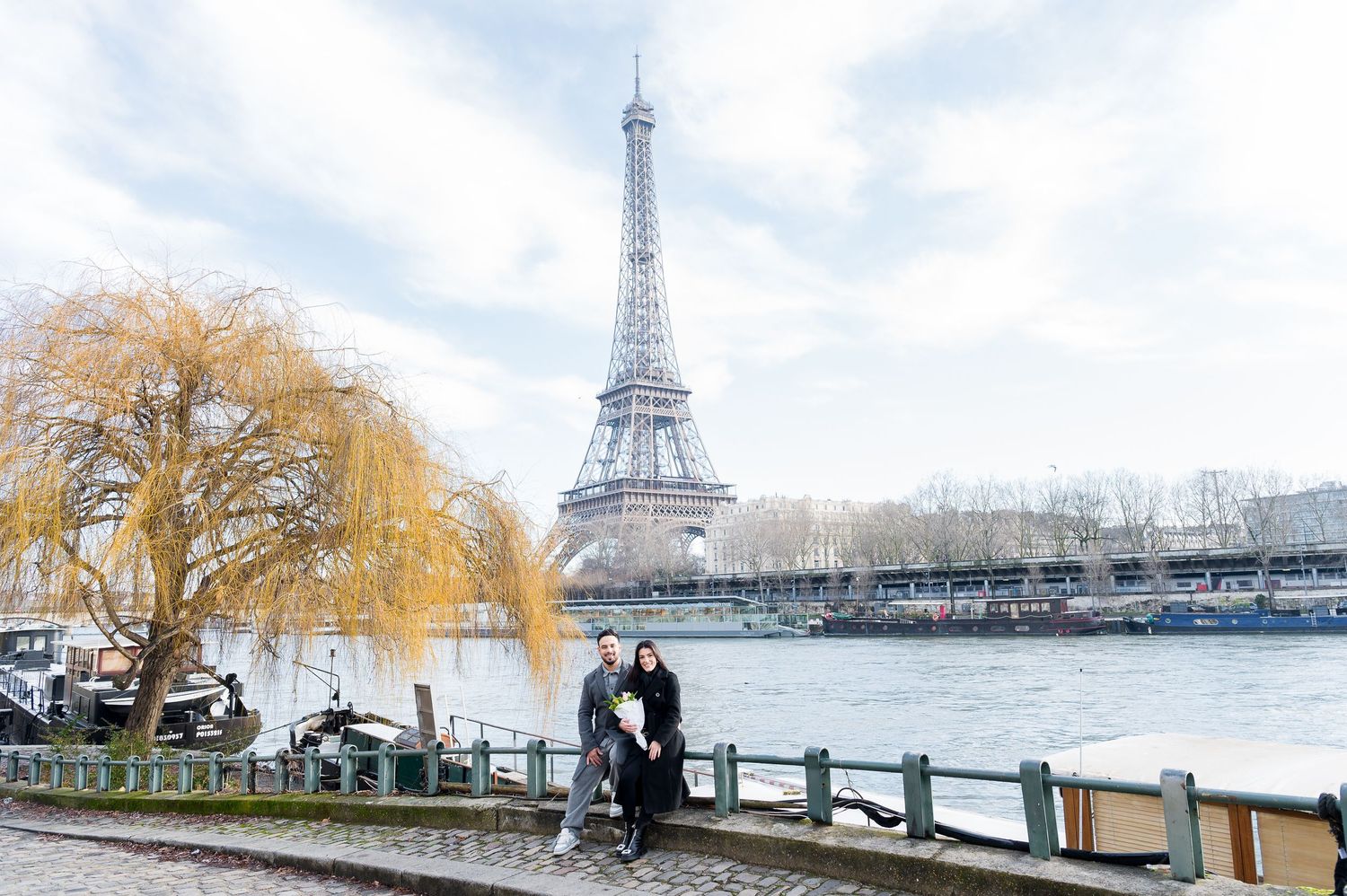 A couple enjoys a romantic winter moment near the Seine with the Eiffel Tower and bare trees in view.