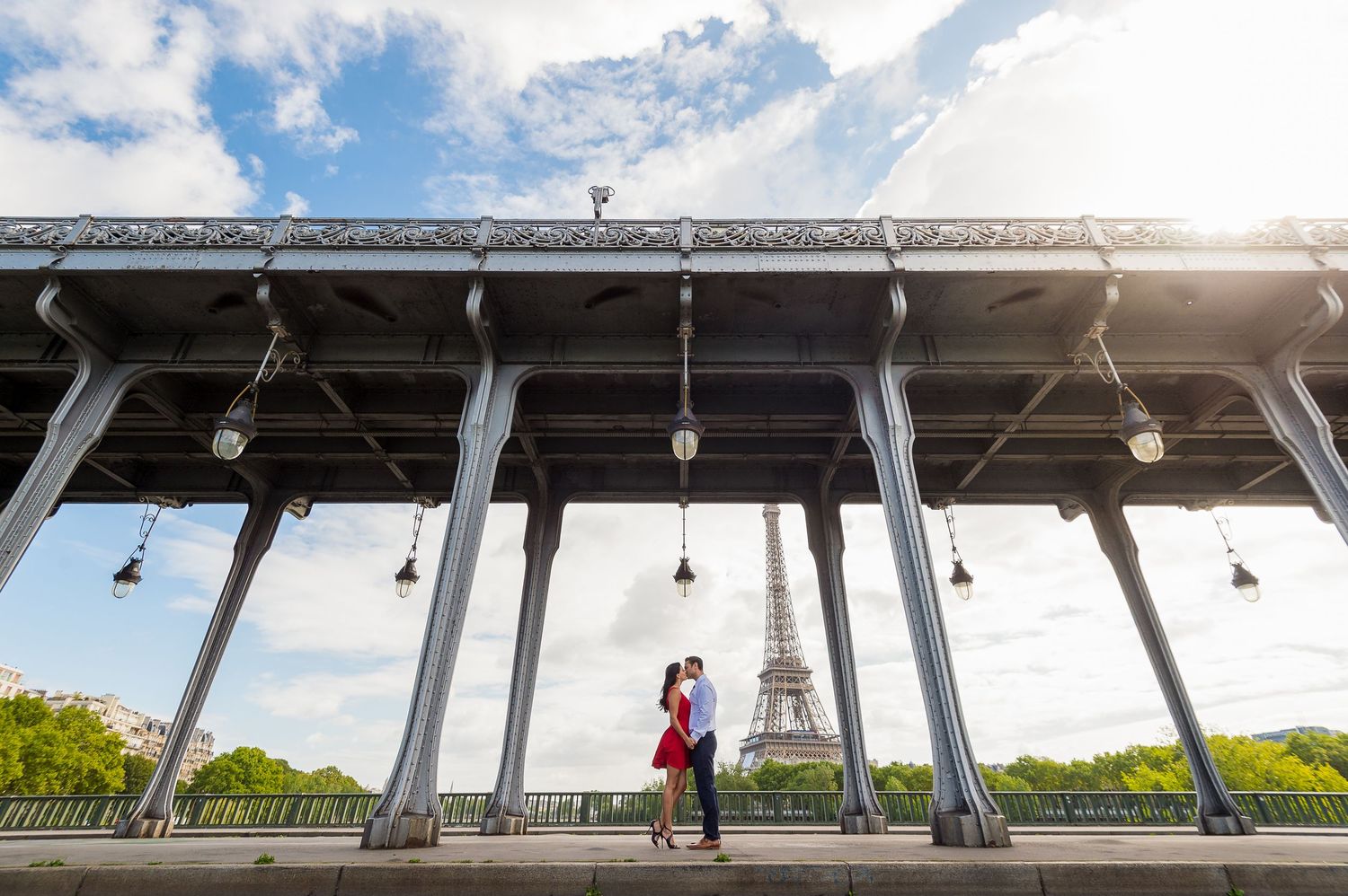 A couple shares a romantic moment under a bridge structure with the Eiffel Tower visible in the distance.