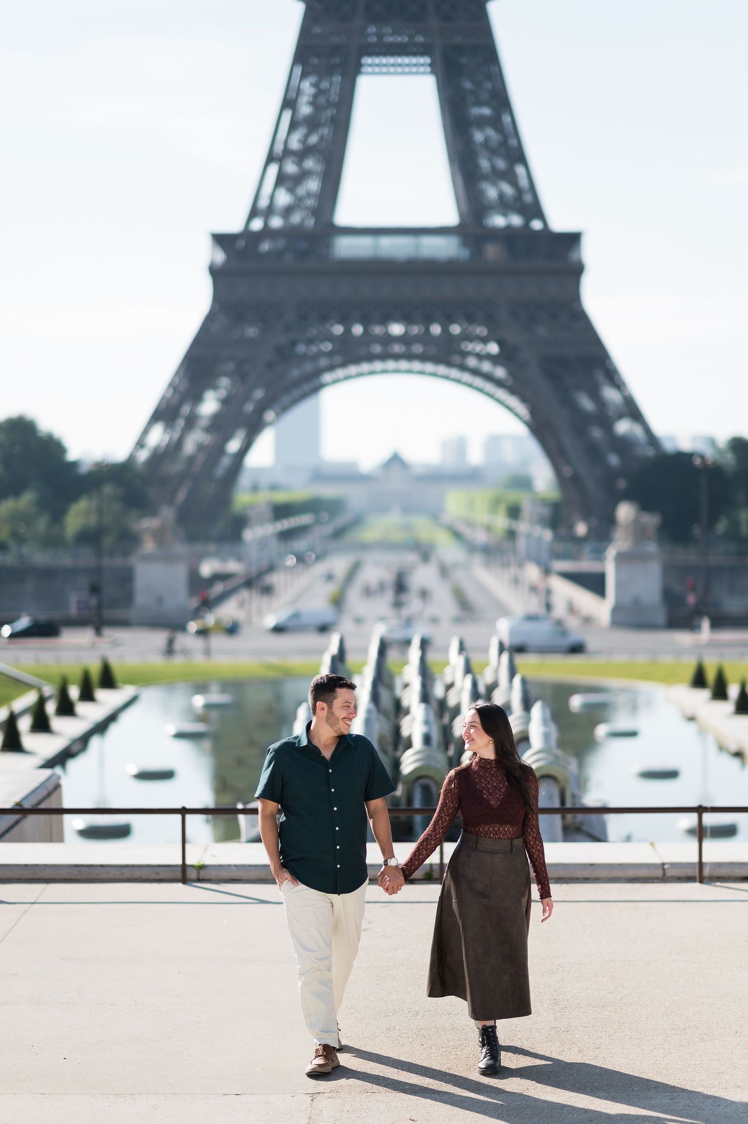 A couple holds hands in front of the Eiffel Tower in Paris, France, with the Champ de Mars in the background.
